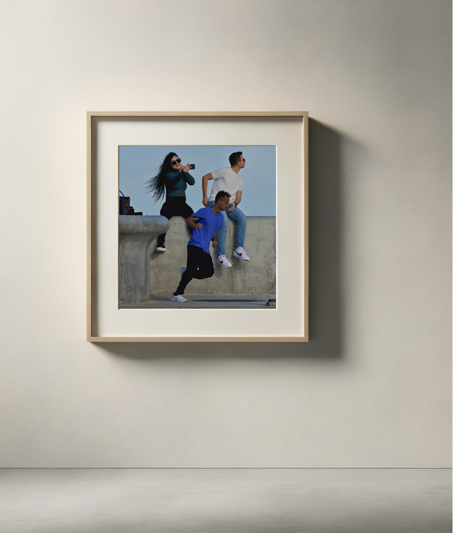 A framed photo on a wall showing three young people near a concrete barrier, two sitting on it and one running, with the girl taking a photo of the scene.