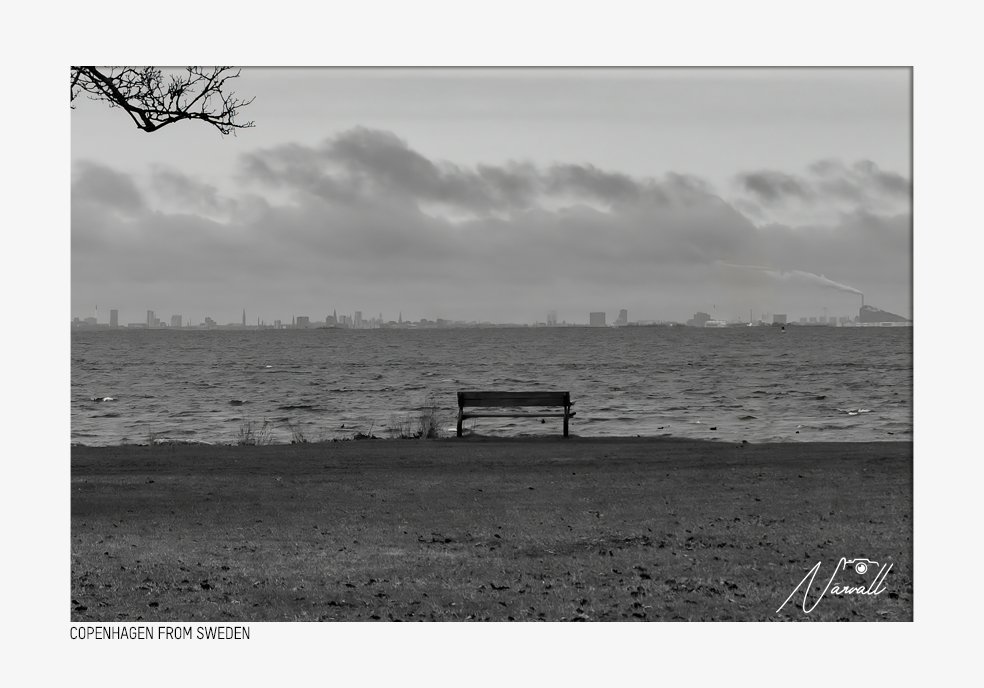 A black and white photo of a beach with a bench facing the water, a city skyline with tall buildings in the distance, and a cloudy sky. The photo has a signature in the lower right corner and the caption "COPENHAGEN FROM SWEDEN" at the bottom.