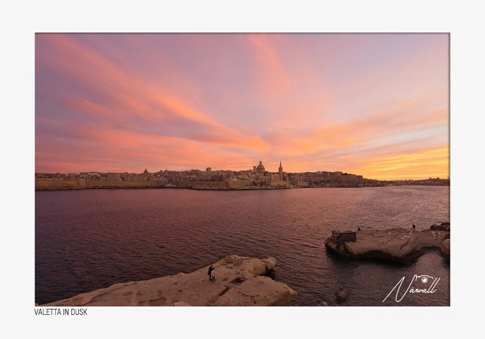 Sunset over a cityscape with historic buildings, including domes and spires, across a body of water with rocky shoreline in the foreground, during dusk with pink and orange clouds in the sky.
