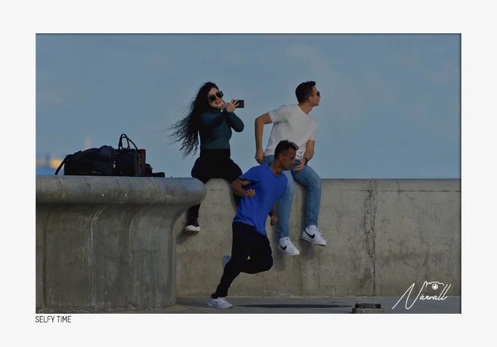 Three young adults on a concrete ledge by the water; one woman taking a selfie and two men sitting and standing nearby, with bags beside them.