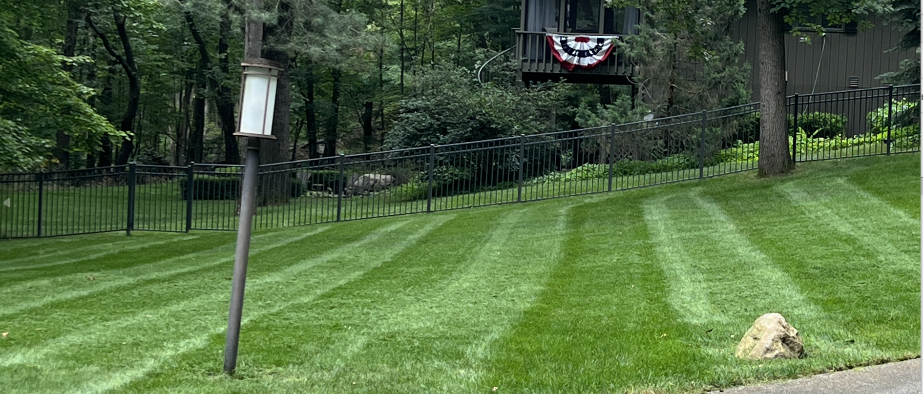 Well-maintained green lawn with a rock and a black metal fence, trees in the background, and a house with a deck decorated with patriotic bunting.