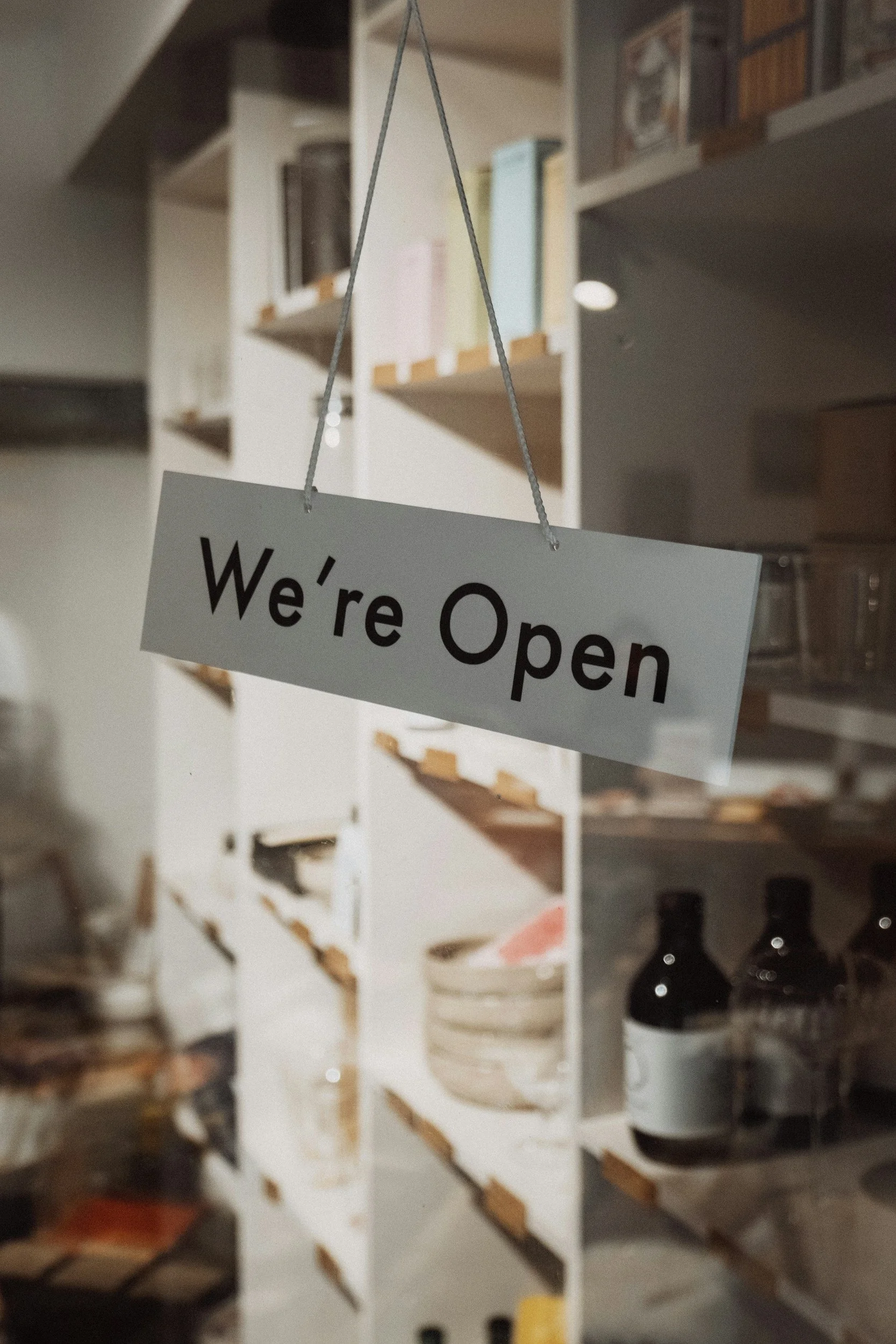 A white 'We're Open' sign hanging with a gray string in front of a blurred background of shelves with books and bottles.