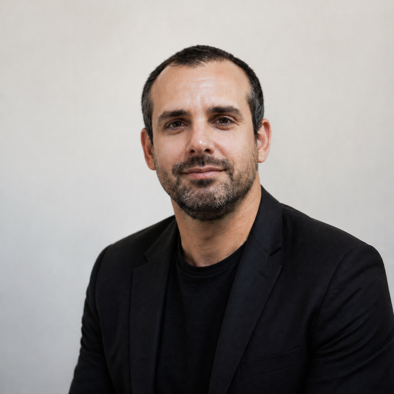 Portrait d'un homme avec barbe, portant un blazer noir, contre un fond blanc, regardant vers l'objectif et souriant légèrement.