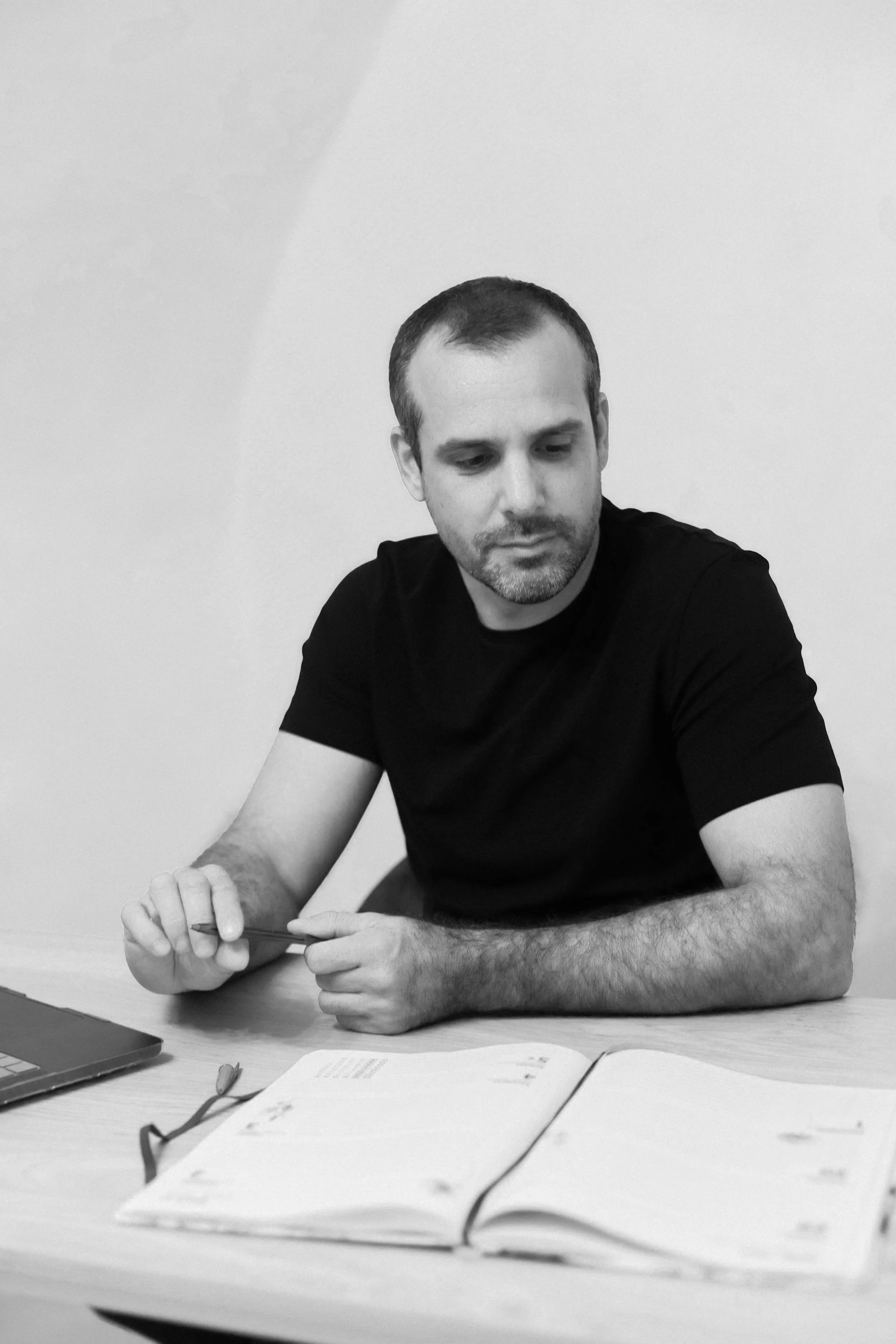 Un homme assis à une table, regardant un livre ouvert avec un ordinateur portable à côté, en noir et blanc.