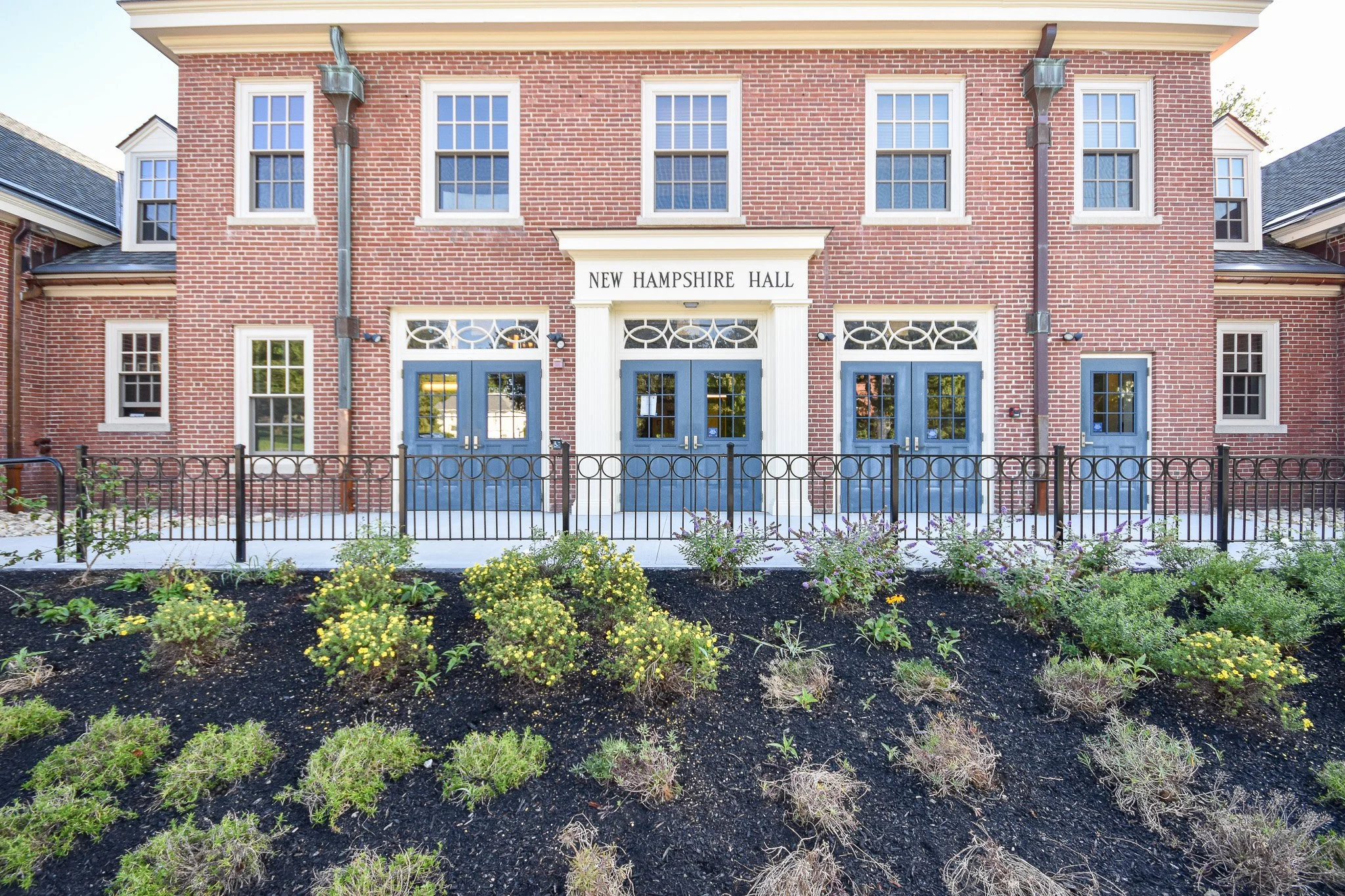 Front view of a two-story red brick building with blue doors and multiple white-framed windows, labeled 'New Hampshire Hall,' with a black metal fence and landscaped flower bed in the foreground.