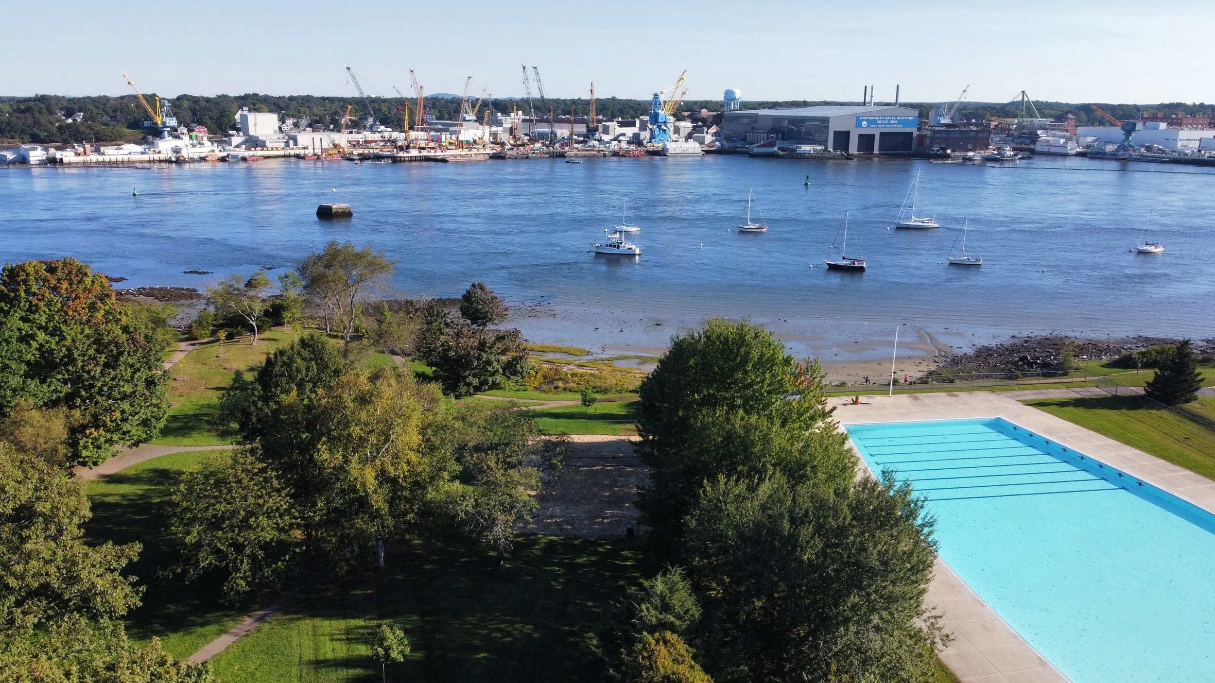 A view of a harbor with sailboats on calm water, green trees in the foreground, and a large industrial shipyard with cranes in the background.