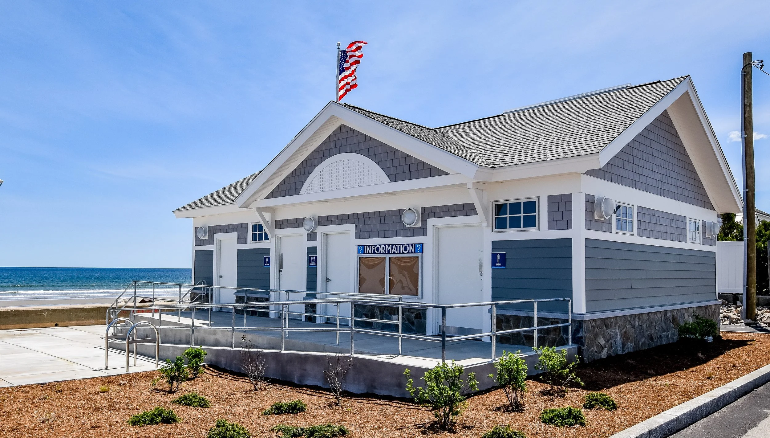 A beachside building with an American flag on top, serving as an information station. The building has gray and white siding, a shingled roof, and signs indicating men's and women's restrooms. It is situated near the ocean, with a clear blue sky overhead and a paved walkway with railings in the foreground.