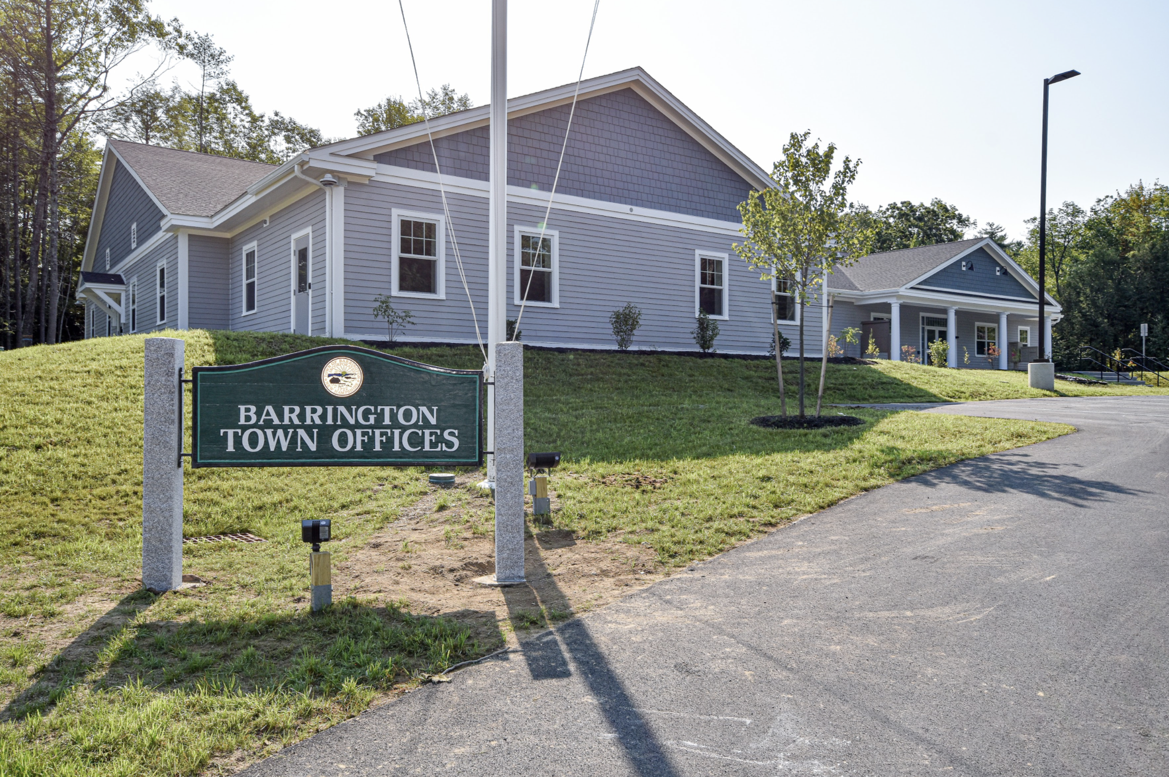The image shows the exterior of Barrington Town Offices building, a two-story structure with gray siding and white trim. There are several windows, a small front porch with steps, and a green sign in front that reads 'Barrington Town Offices.' The surrounding area has a well-maintained grassy lawn with trees, and a sidewalk runs past the building. The weather is clear and sunny.