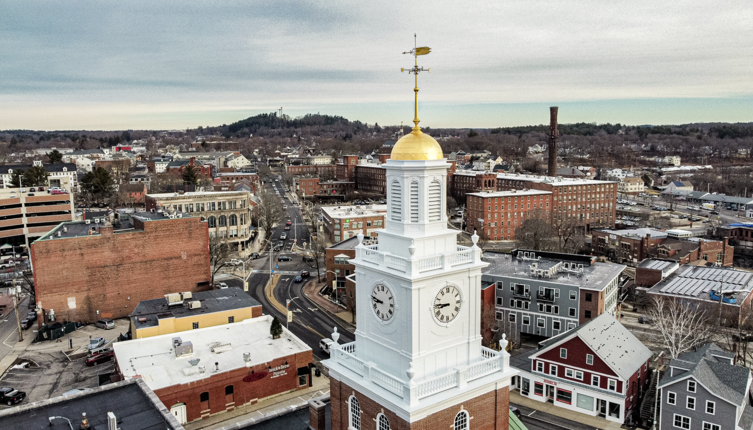 A cityscape featuring a white clock tower with a gold dome and weather vane on top, surrounded by various commercial and residential buildings, streets, cars, and a hilly background under a partly cloudy sky.