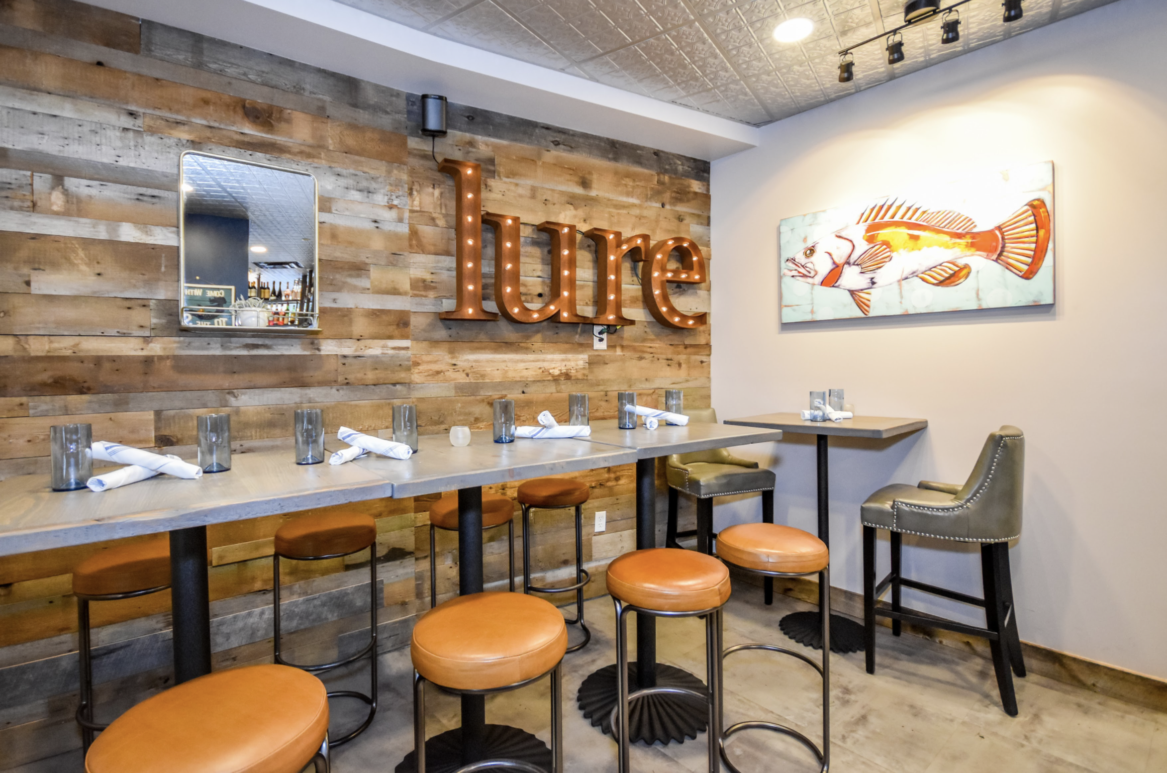 Interior of a restaurant with a wooden wall, a neon sign that reads 'ure', a fish painting on the wall, and tables set with glasses and napkins.