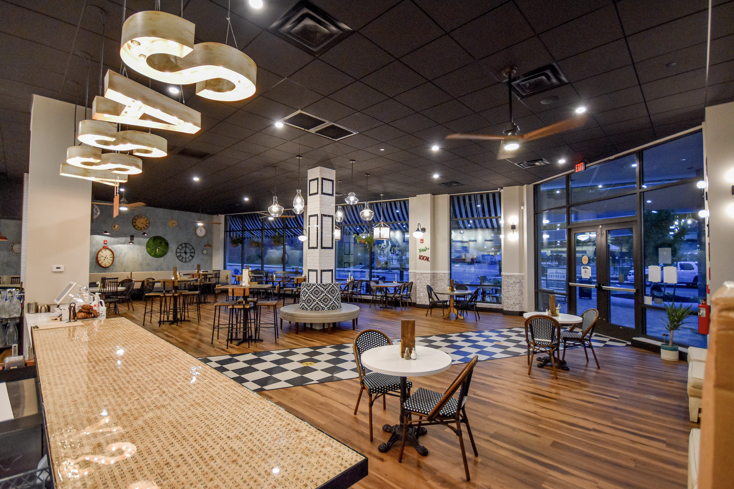 Interior of a modern restaurant with wooden floors, various tables and chairs, large windows, decorative hanging lights, and ceiling fans. The decor includes wall clocks and plants.