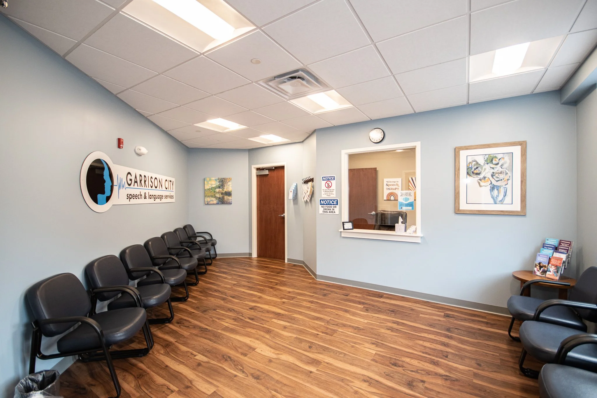 Waiting room with black chairs, a wall sign for Garrison City Speech & Language Services, framed artwork, brochures, and a reception window.