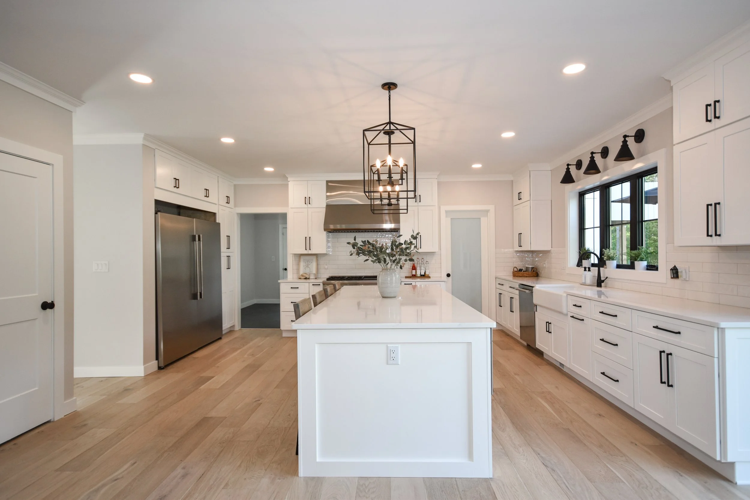 Bright modern kitchen with white cabinets, stainless steel refrigerator, black hardware, large island with white countertop, black light fixture, wood flooring, and windows with greenery outside.