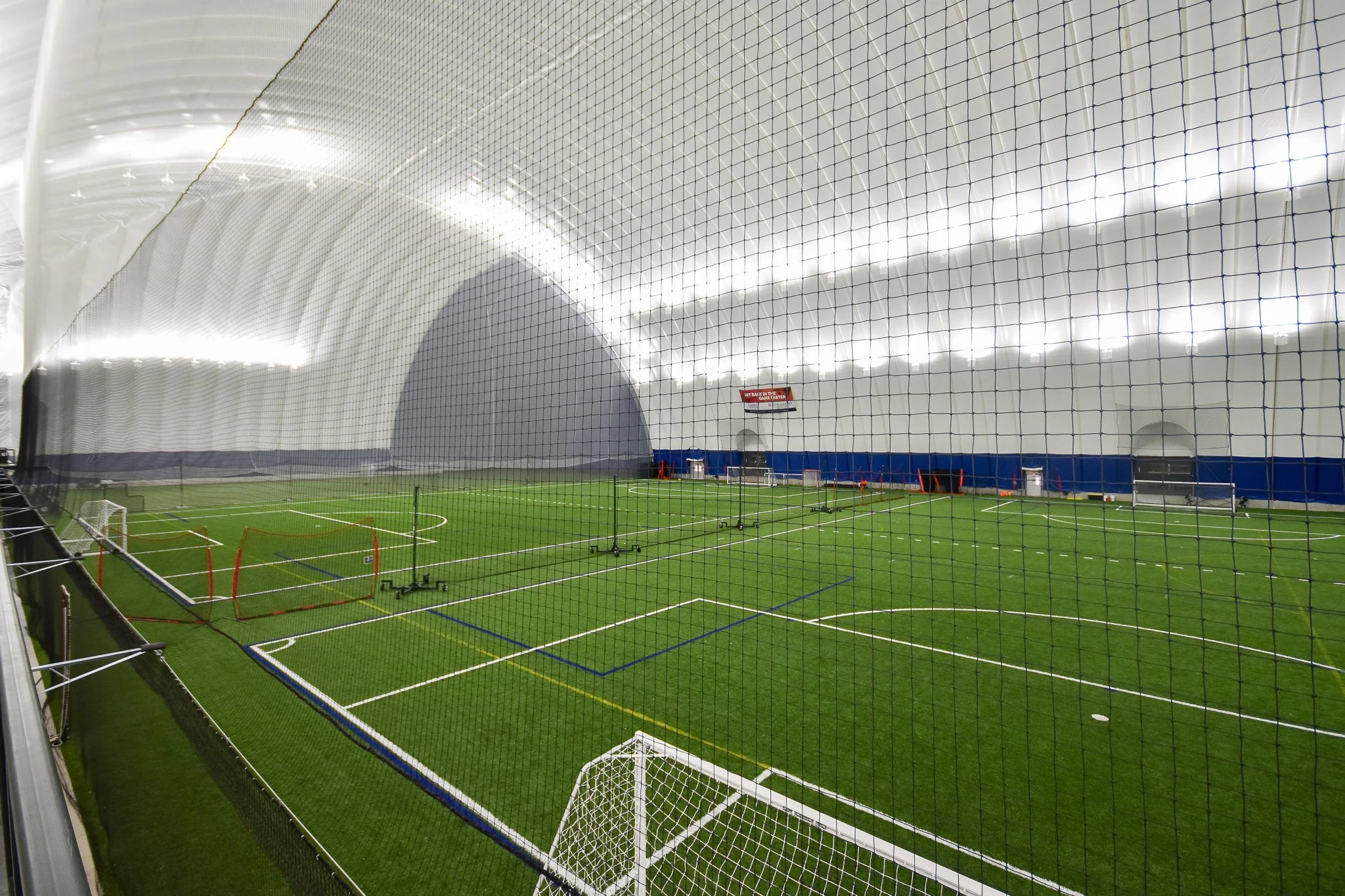 Indoor soccer field with artificial turf, surrounded by a large net, with goals at each end and a sign reading 'Get Back in the Game Laster'