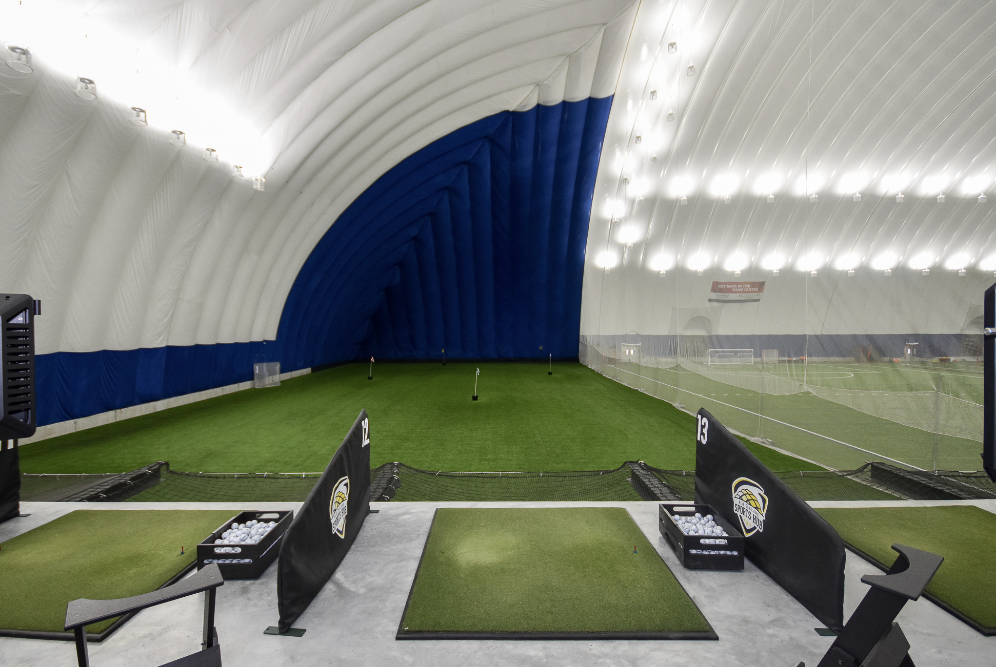 Indoor baseball training facility with artificial turf, pitching targets, and golf simulation nets, inside a large white inflated dome with bright lighting.