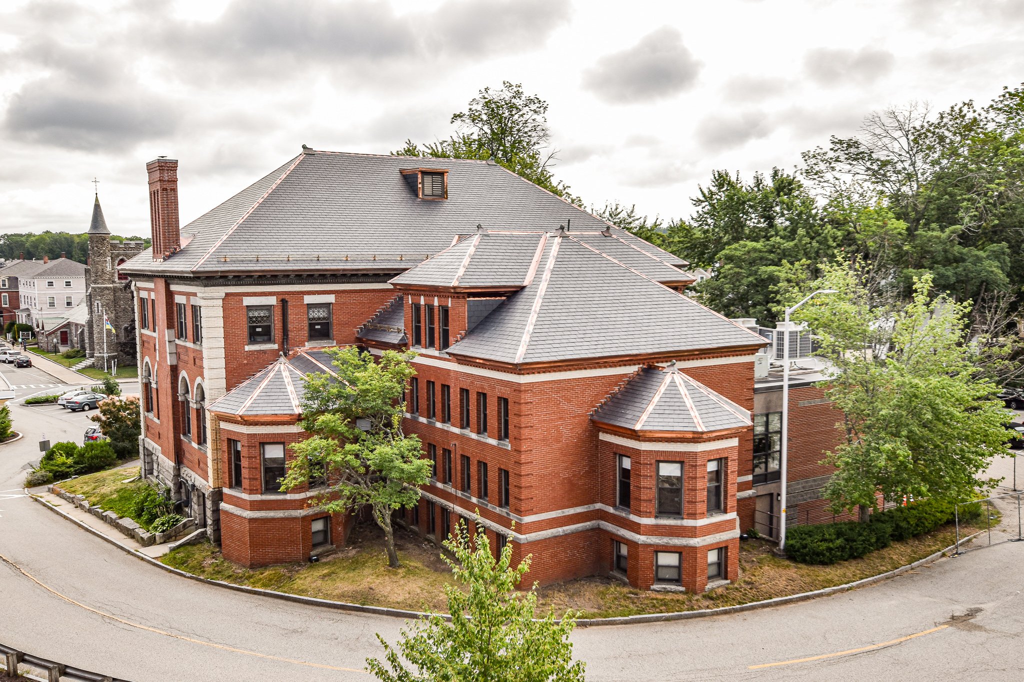 A large red brick building with multiple rooflines and a chimney, surrounded by trees and a curved street, under a cloudy sky.