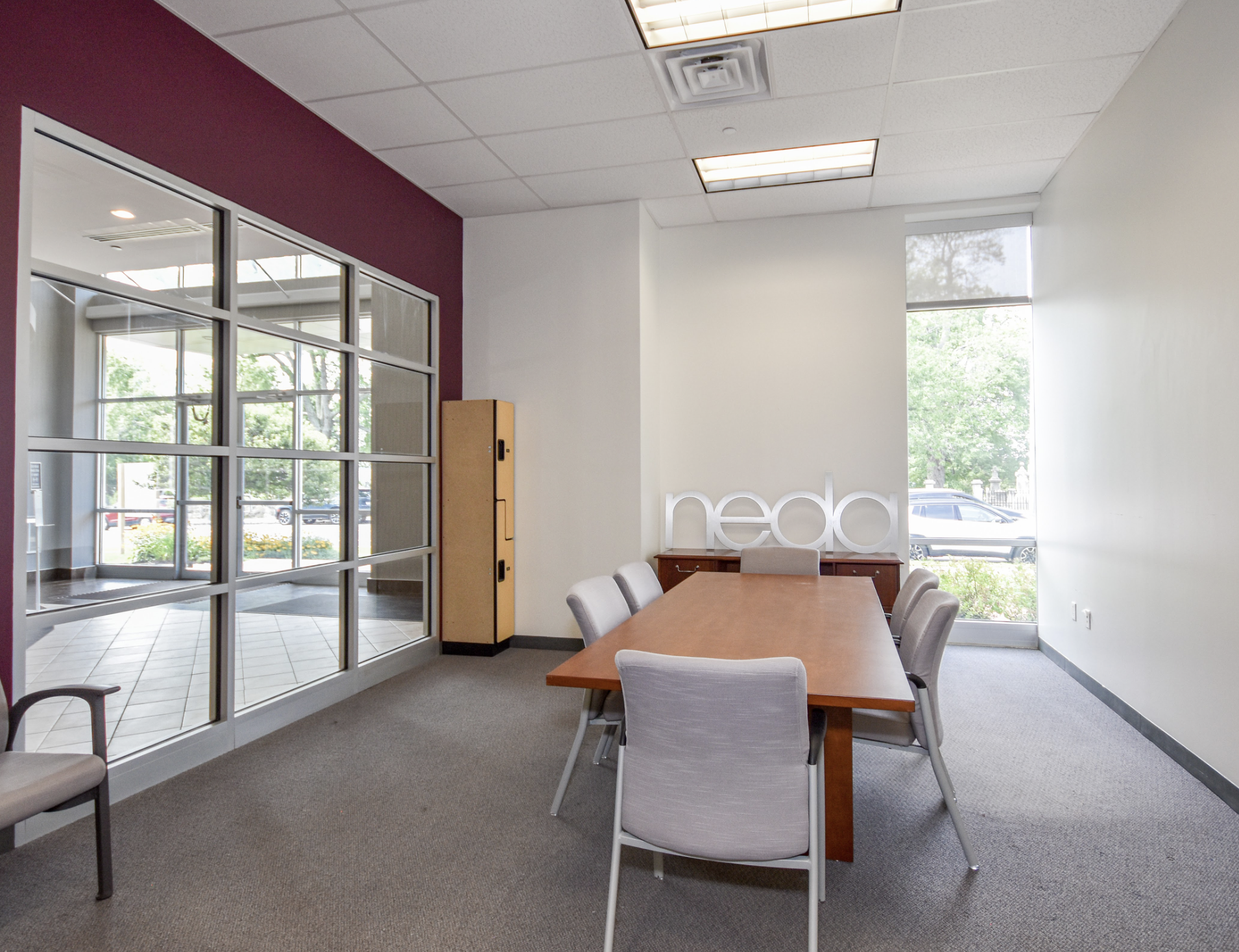 Meeting room with wooden table, gray chairs, large window with trees outside, and sign that says 'neda' on the wall.
