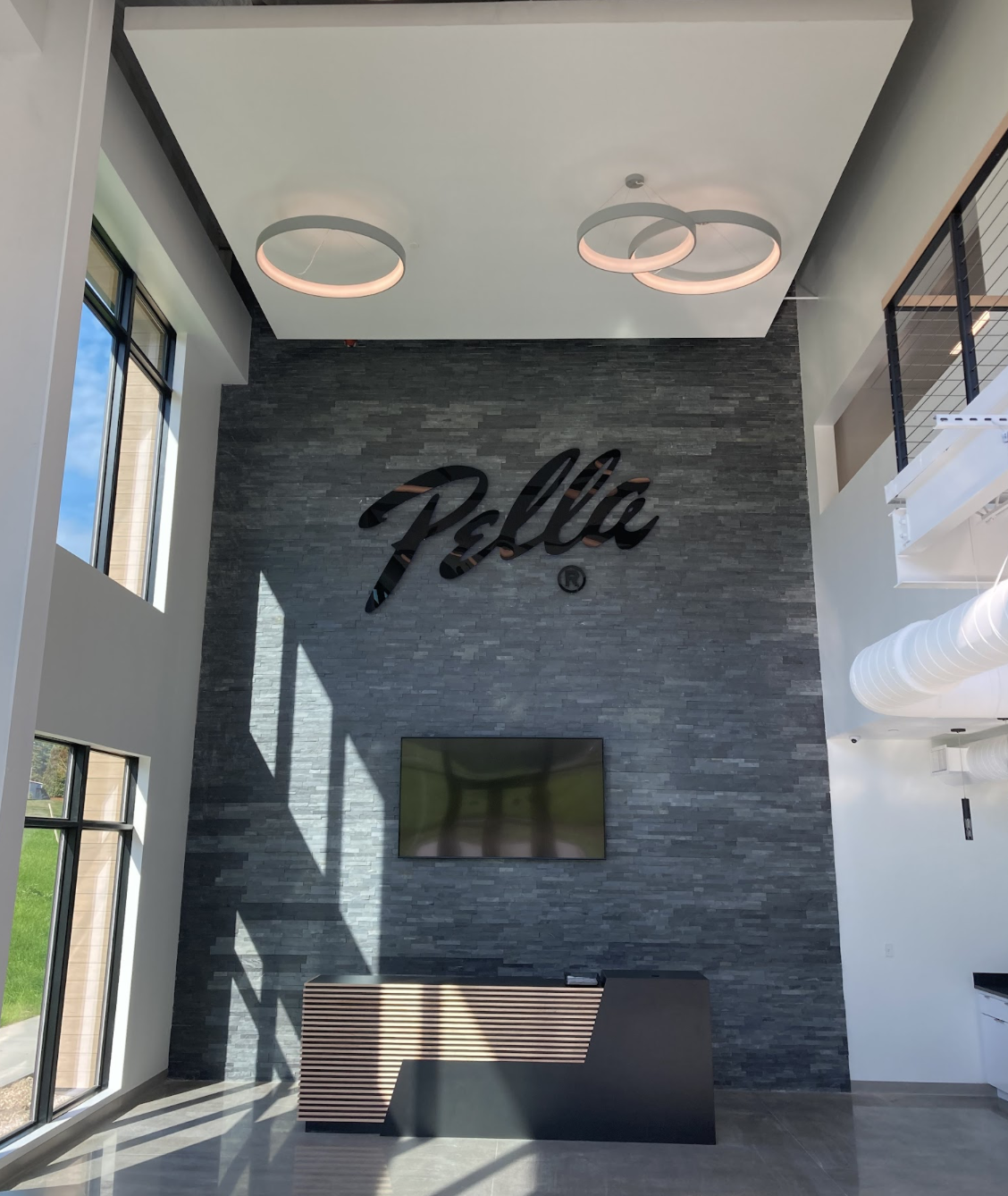 Modern interior lobby with a large black 'Pella' logo on a dark gray stone accent wall, a flat screen TV, and decorative modern ceiling lights with circular rings, next to large windows letting in natural light.