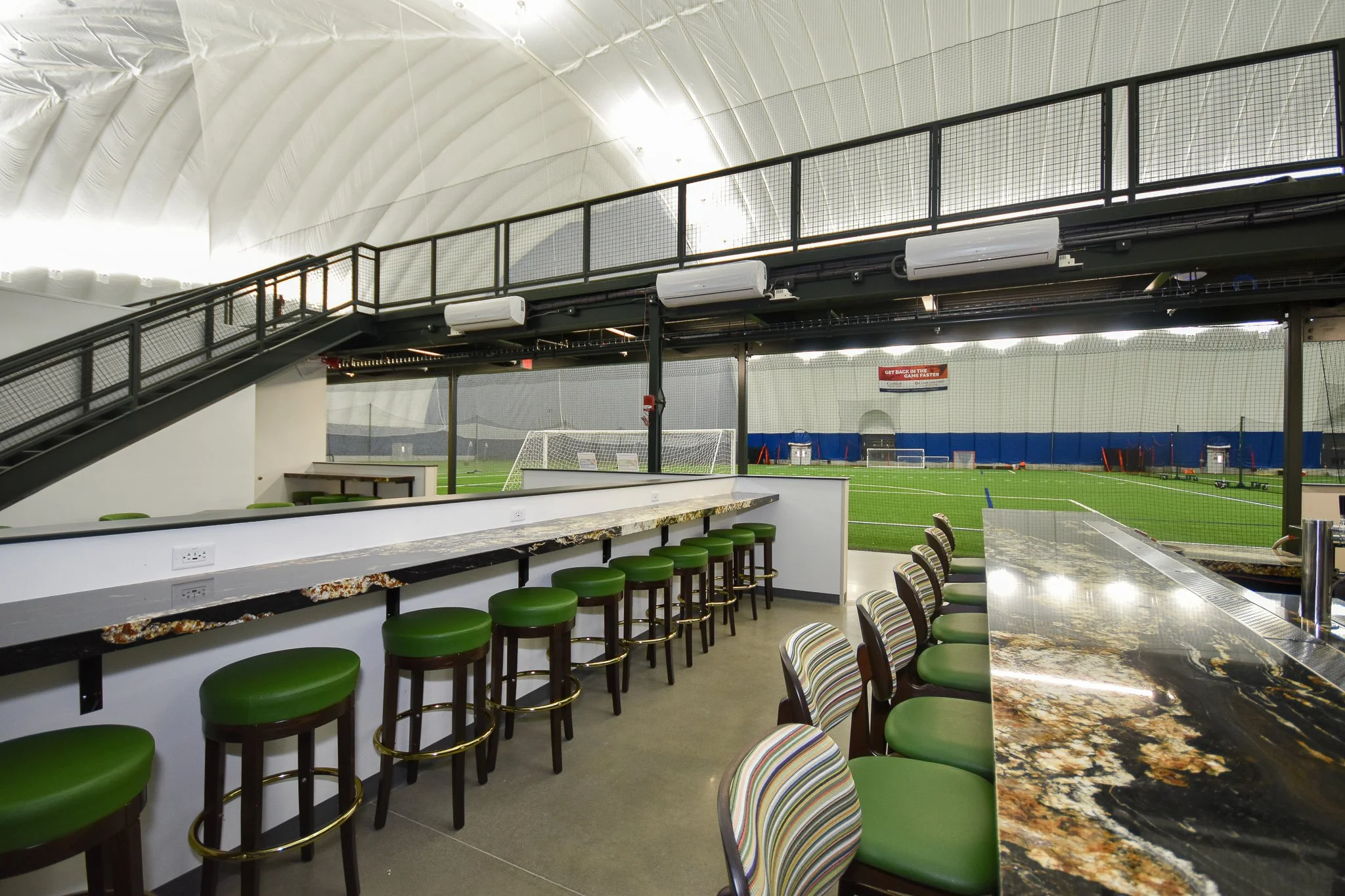 Indoor sports facility with a soccer field visible through large windows, a staircase with black railing leading to a second level, and a seating area with green barstools and a marble countertop.