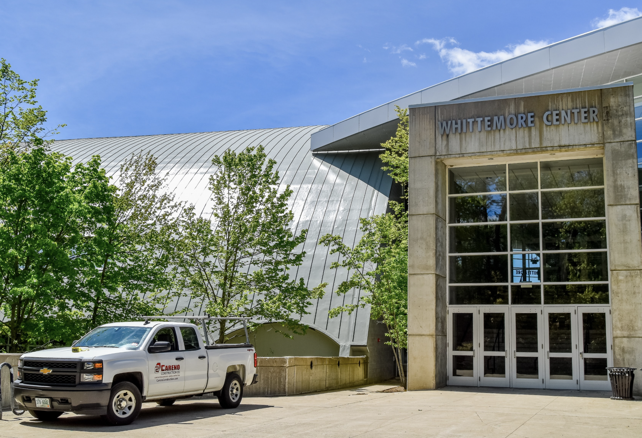 Exterior view of Whittmore Center with trees, a white Chevrolet pickup truck parked in front, and a blue sky with some clouds.