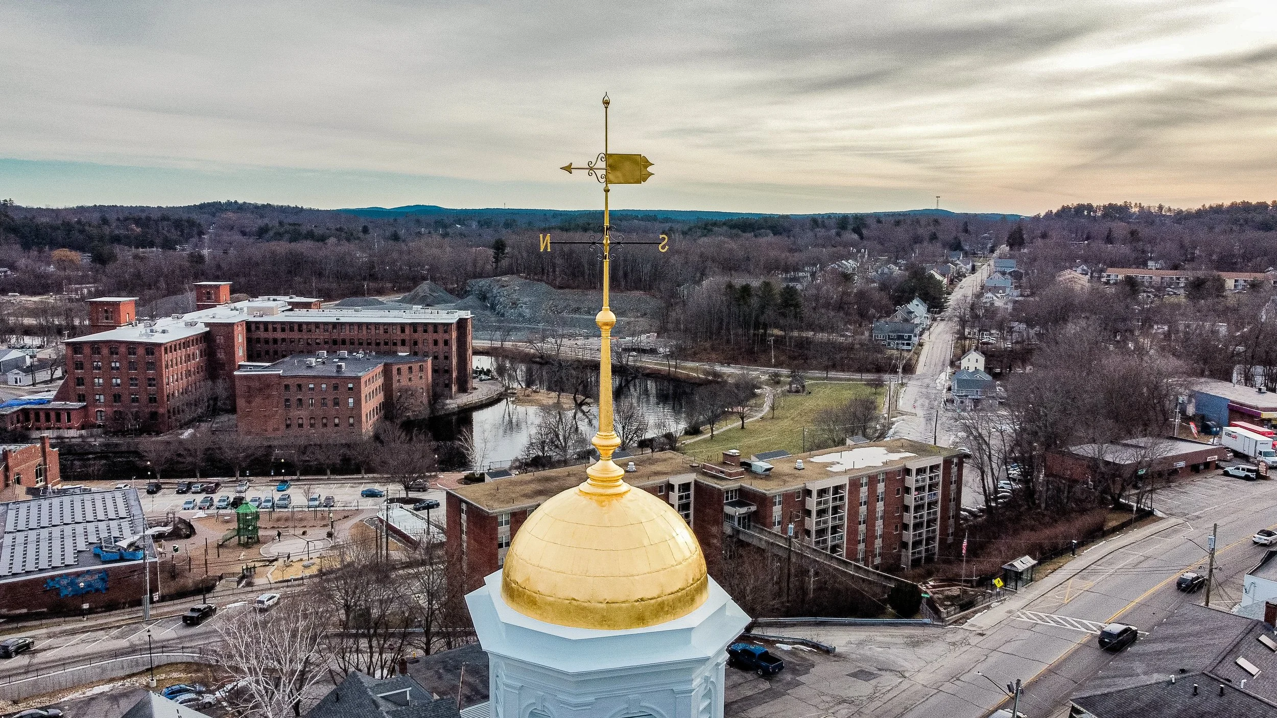 View of a town with a mix of historic and modern buildings, a pond, and a hilly landscape in the background, as seen from a high vantage point with a golden dome and weather vane in the foreground.