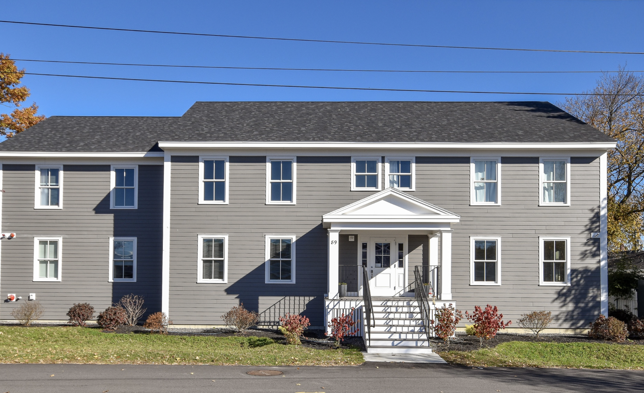 A gray, three-story residential building with white trim and a gabled roof, facing a street with a grassy front yard and bushes. There is a small porch with stairs leading to the front entrance, and power lines are visible above. The sky is clear and blue, with some trees showing fall foliage in the background.