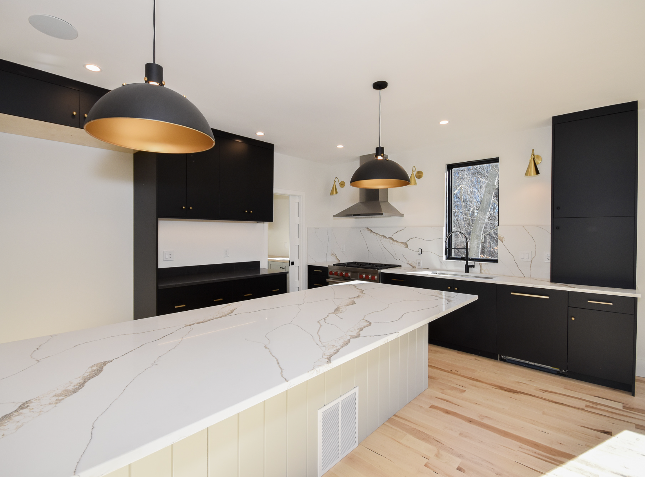 Modern kitchen with black cabinets, white marble countertops, and gold accents. Pendant lights hang over the island, and a window shows a snowy outdoor scene.