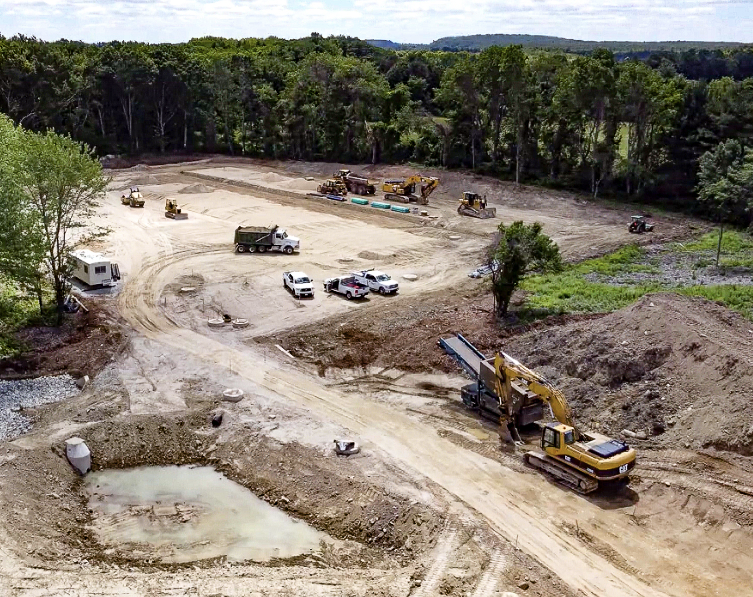 Construction site with heavy machinery, trucks, and vehicles working on land clearing or grading, surrounded by trees and forest in the background.
