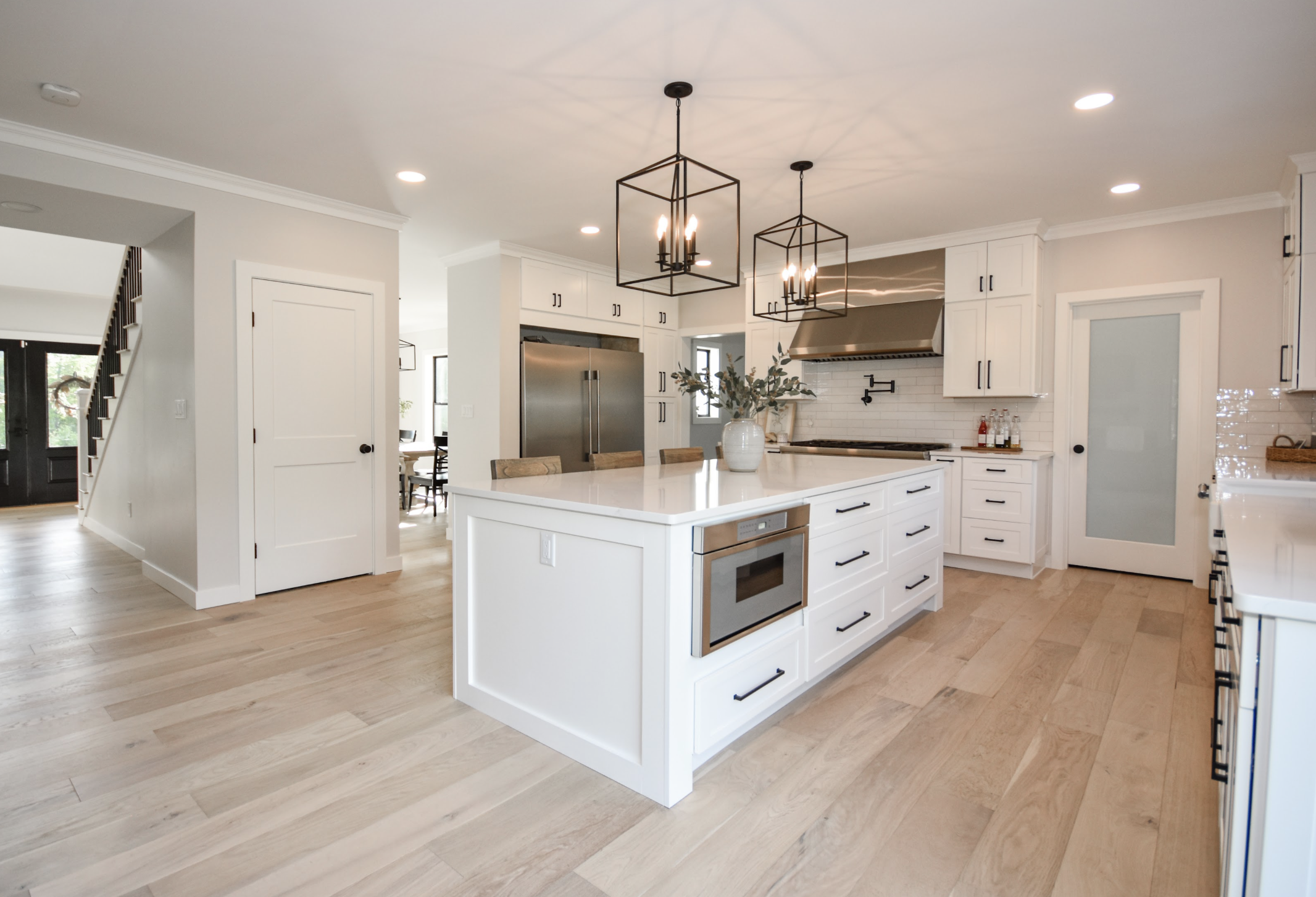 Modern kitchen with white cabinets, a large kitchen island with a vase of greenery, stainless steel appliances, and black pendant lights over the island. Light wood flooring and a white brick backsplash visible.