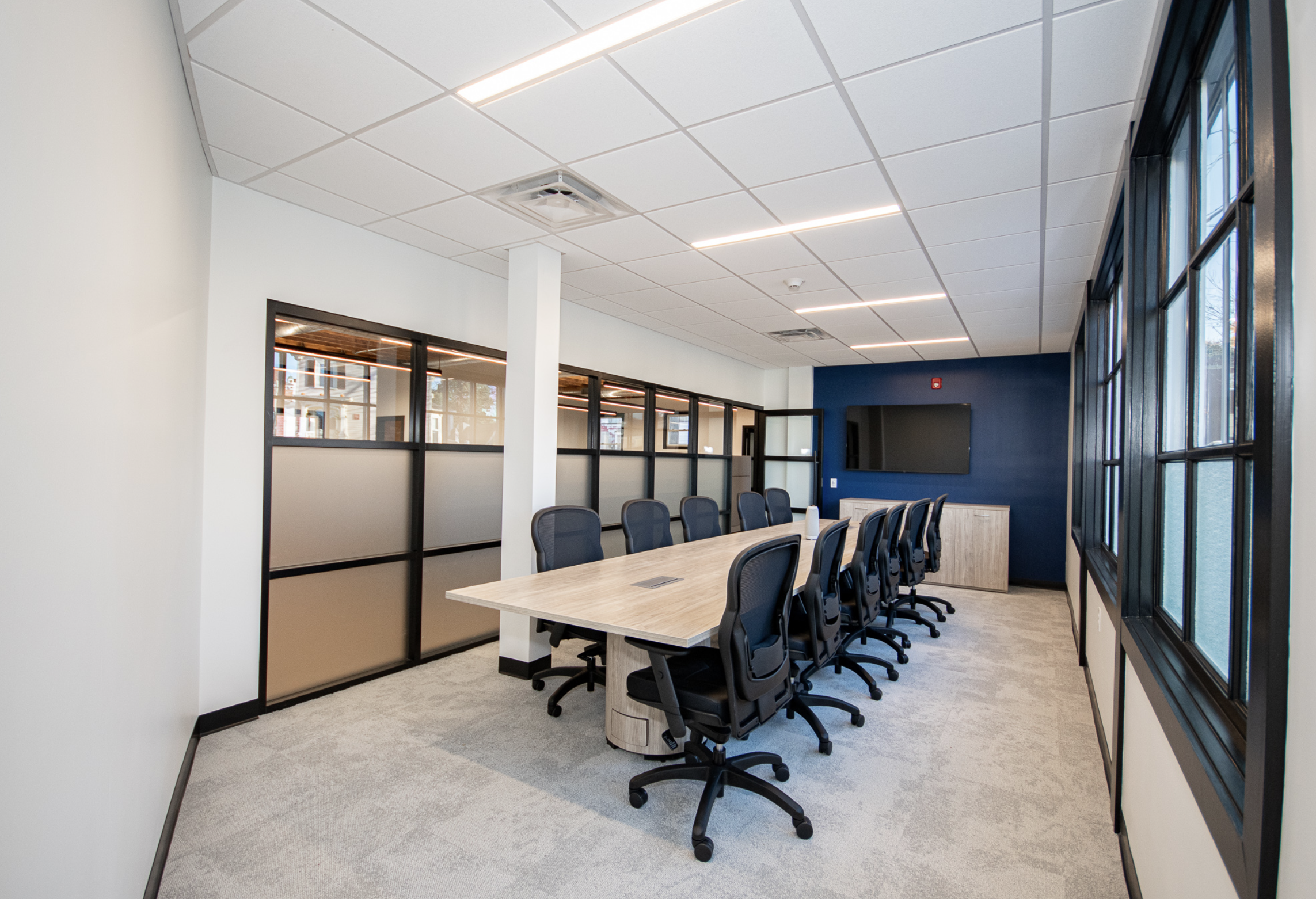 Modern conference room with a long table, black chairs, large windows, blue accent wall, white walls, and ceiling with recessed lighting.