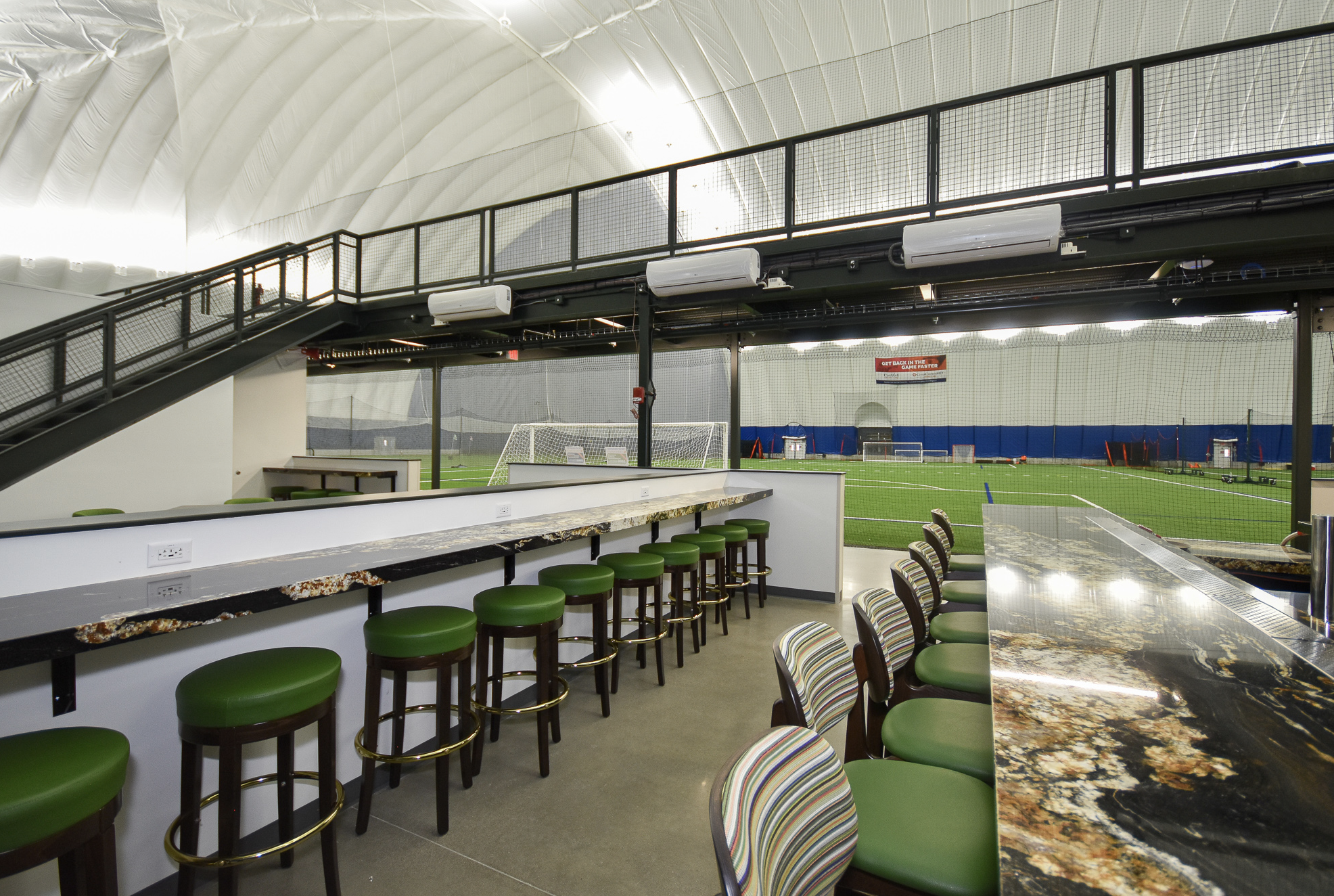 Indoor sports facility with artificial turf, soccer goal, and sports equipment. Inside viewing area with bar seating and marble-top counter.