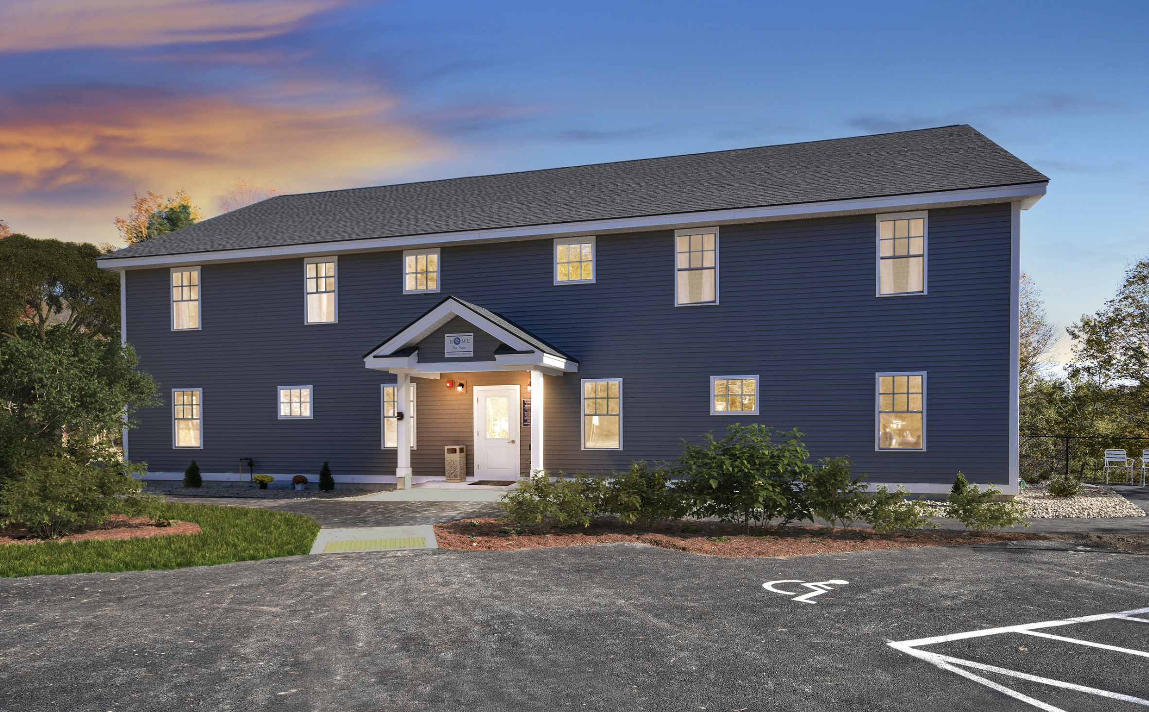A modern multi-story house with blue siding, white trim, and a dark gray roof at sunset. There is a small covered porch, a landscaped front yard, and a parking lot with a designated handicapped parking space in the foreground.