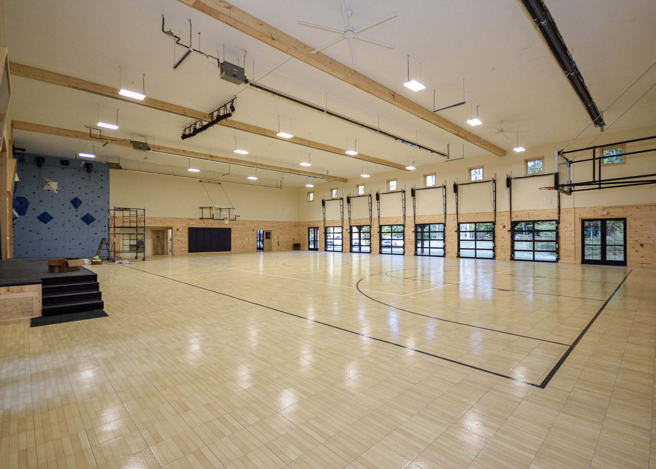 Empty indoor gymnasium with wooden walls, high ceiling with hanging lights, basketball hoops, climbing wall, and large windows.