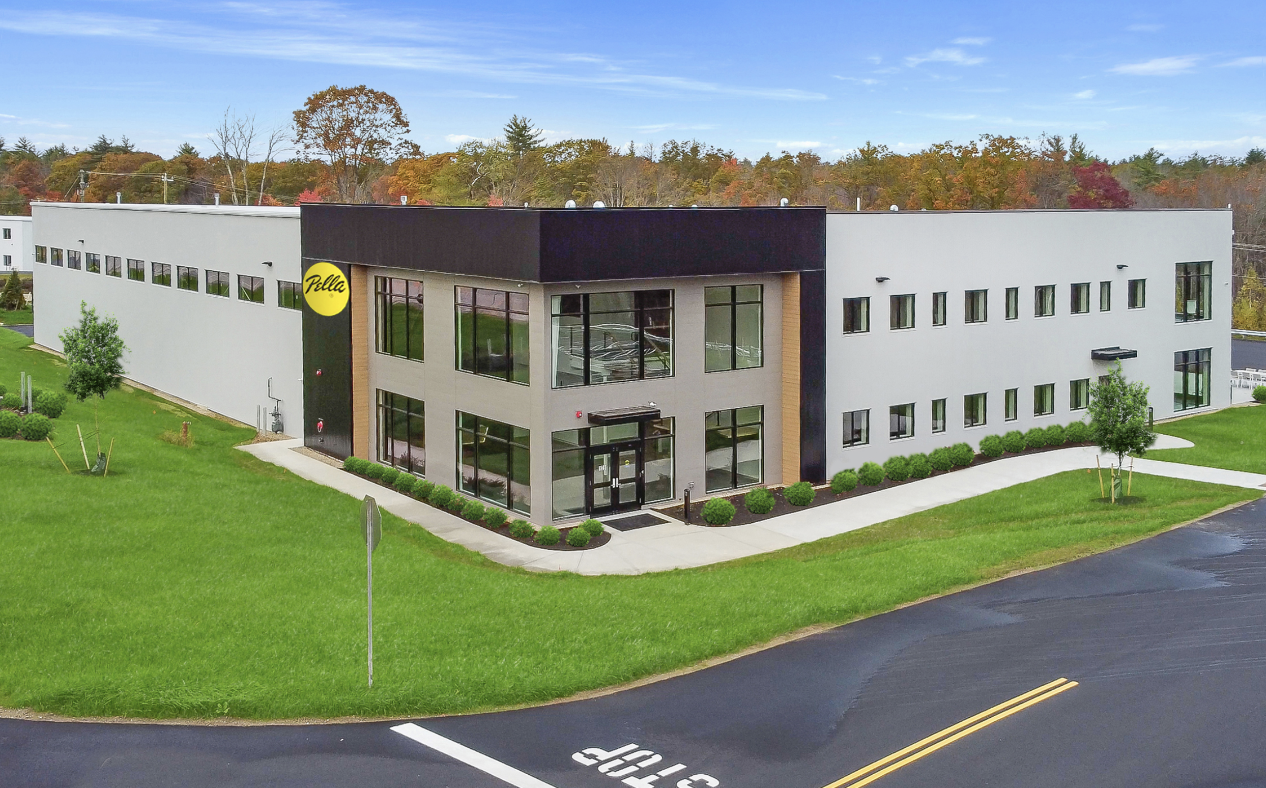 Modern two-story commercial building with large windows, a black and beige exterior, and a yellow circular logo that says 'Pella.' The building is surrounded by well-maintained green grass, trees, and a sidewalk.