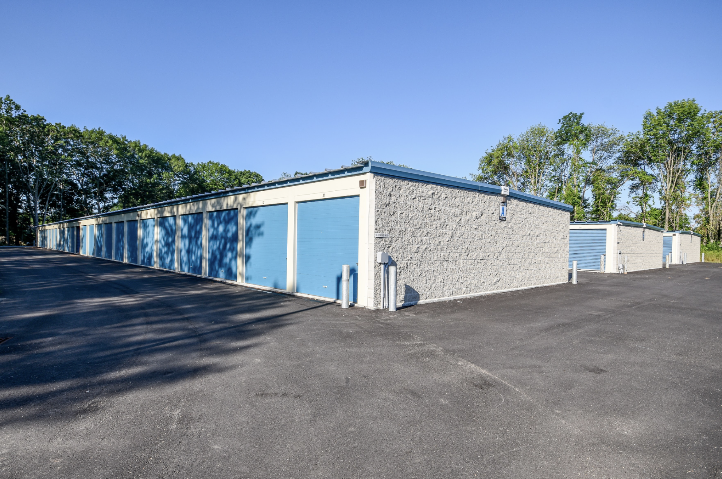 A row of storage units with blue doors, surrounded by trees and a clear blue sky.