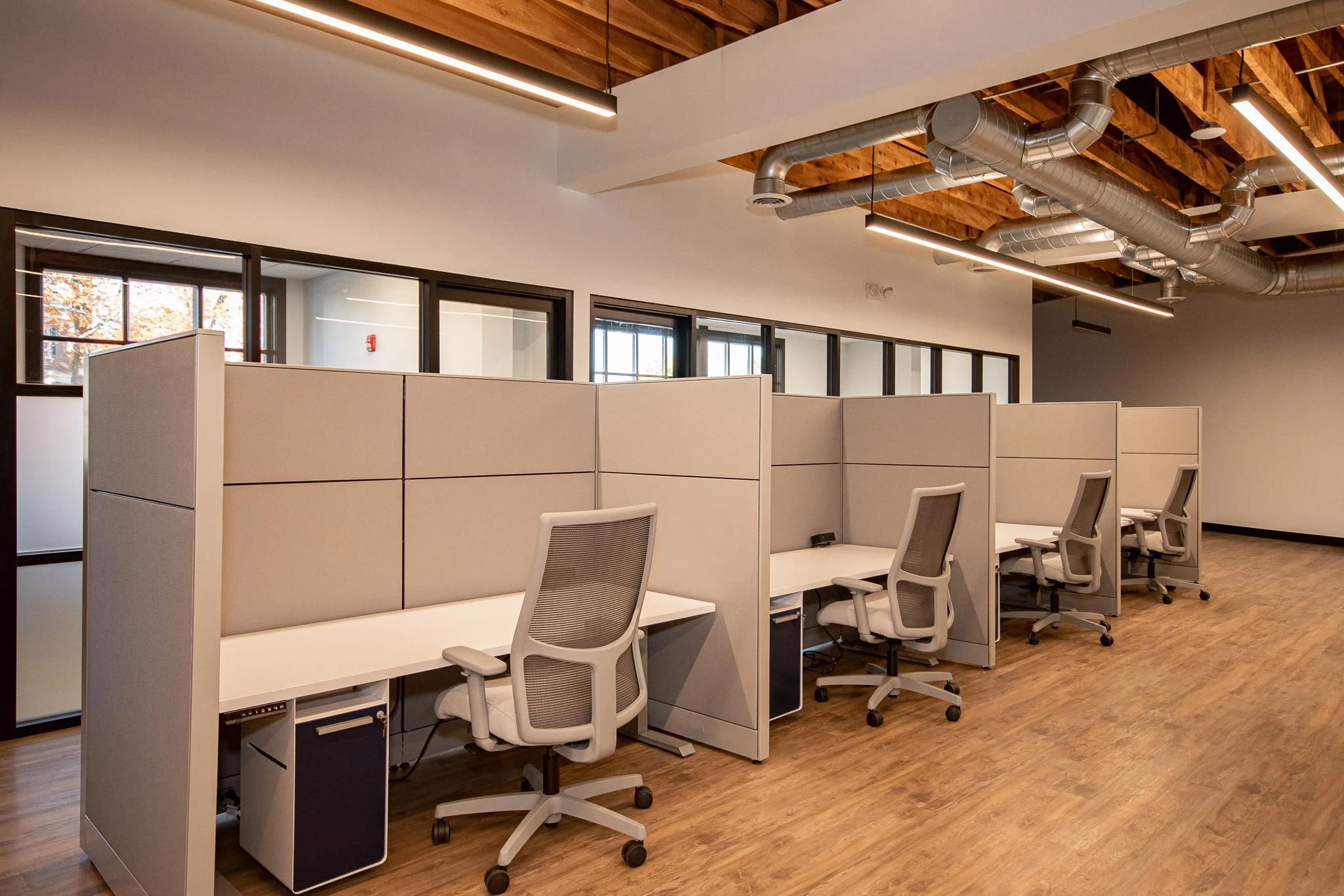 Office cubicles with ergonomic chairs in an open-plan workspace, large windows, and exposed ceiling ducts with modern lighting.