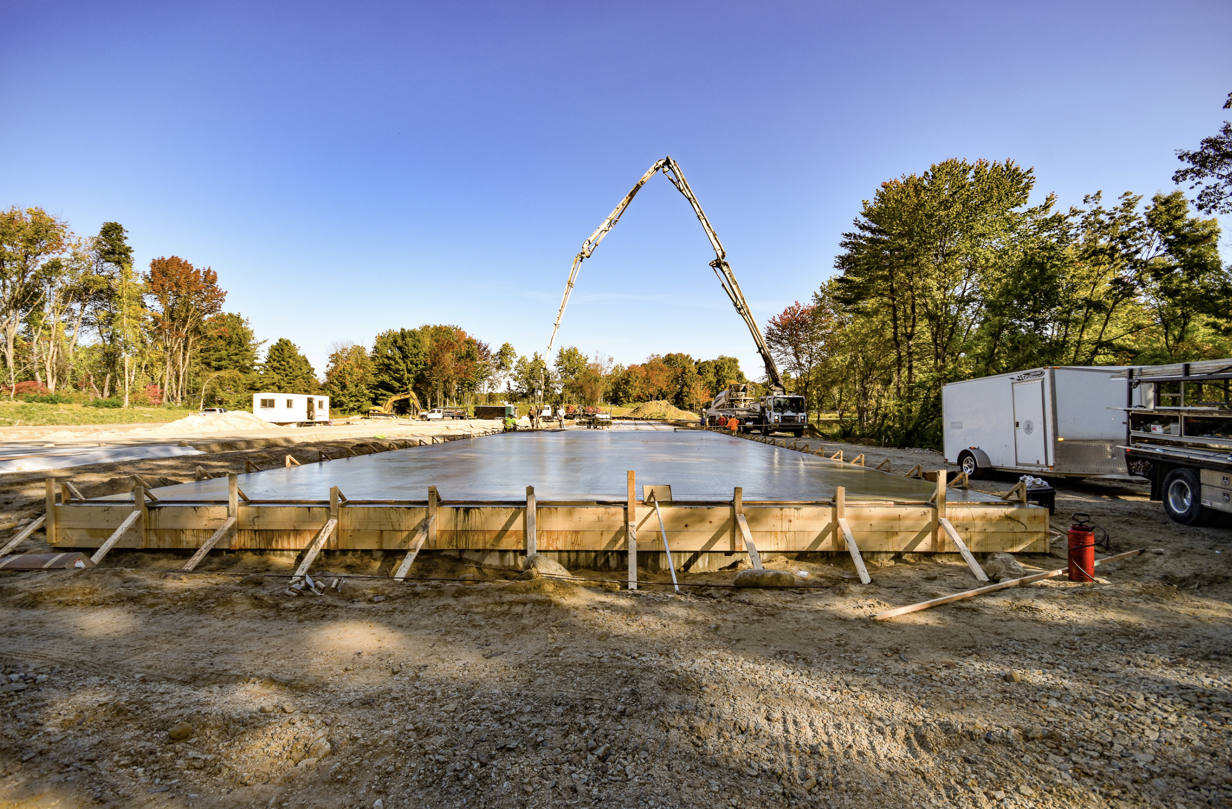 Construction site with a freshly poured concrete slab, surrounded by trees with autumn foliage, construction vehicles, and workers present.