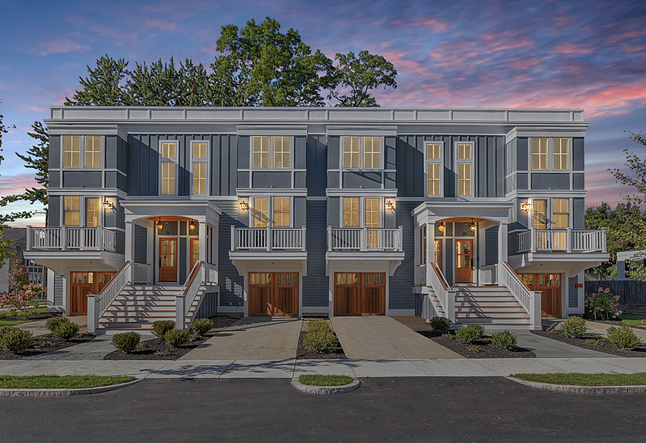 Front view of a modern, multi-story residential building with blue siding, white trim, and steps leading to front doors, set against a sky with pink and purple clouds at sunset.