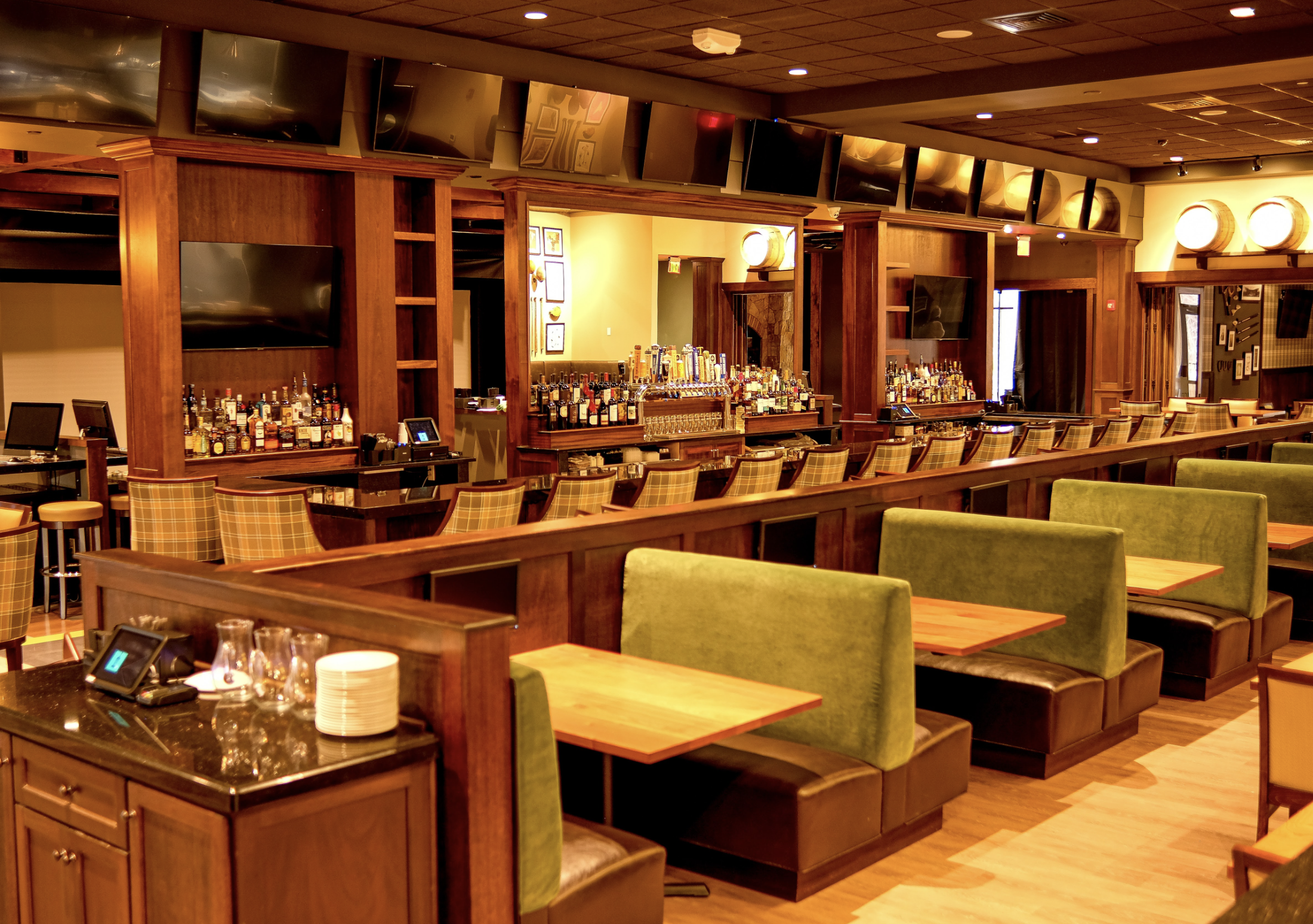 Interior view of a bar with multiple flat-screen televisions mounted above wooden shelves holding liquor bottles, and a bar counter with cash registers and bar stools.