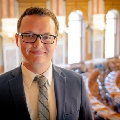 A man in a suit and tie, wearing glasses, smiling in a well-lit room with large windows and wooden accents.