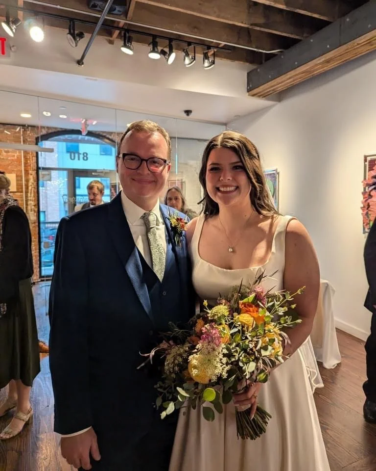 A newlywed couple smiling and posing together indoors at a wedding reception. The groom is wearing glasses, a dark suit, white shirt, and tie, while the bride is in a sleeveless white dress holding a colorful bouquet of flowers.
