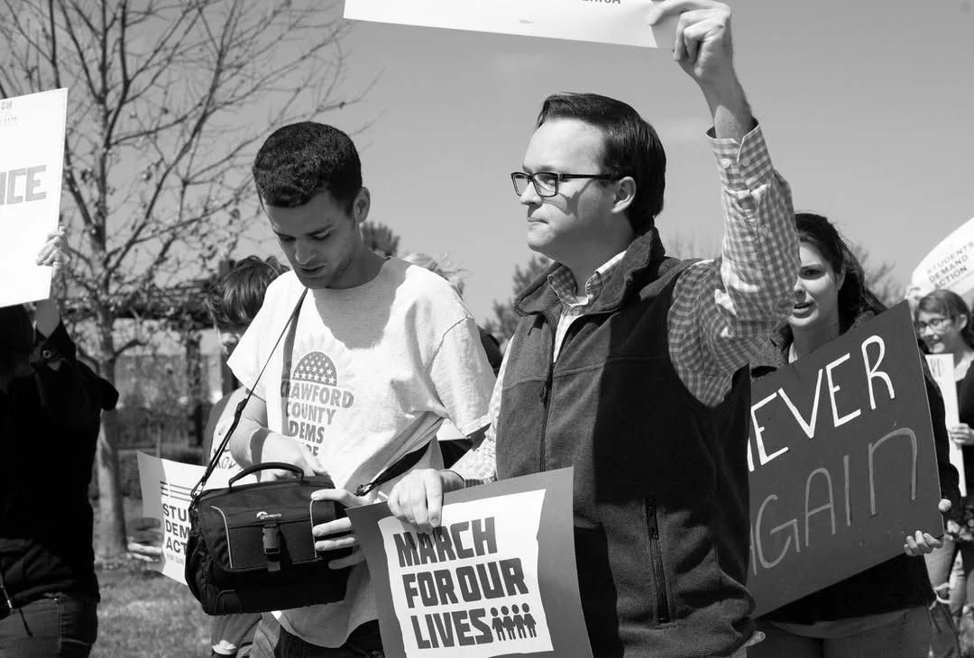 Group of people participating in a march or protest, holding signs with messages like 'March for our lives' and 'Never again.' The scene takes place outdoors with trees in the background.