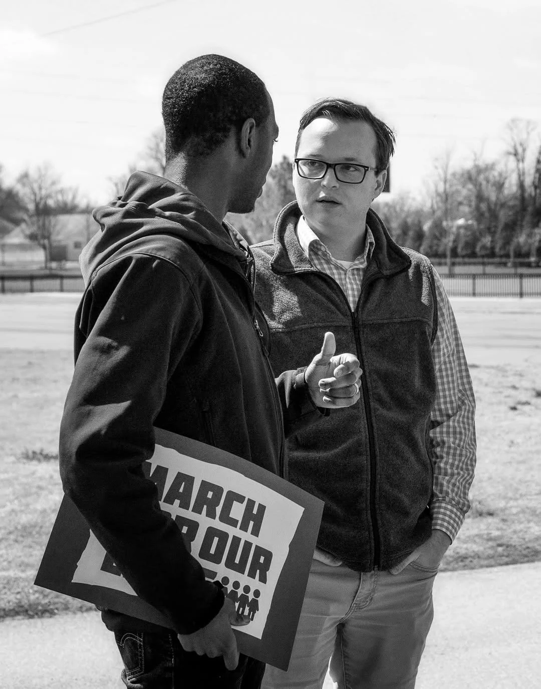 Two men engaged in a conversation outdoors during the day, one holding a sign that reads "March for Our" with an illustration of people beneath the text.