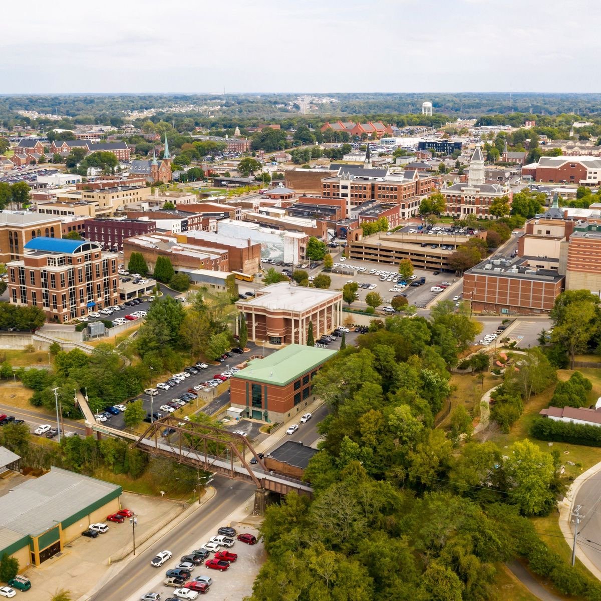 An aerial view of a downtown cityscape with various buildings, parking lots, trees, and streets. The skyline features churches, a water tower, and a mix of modern and historic architecture.