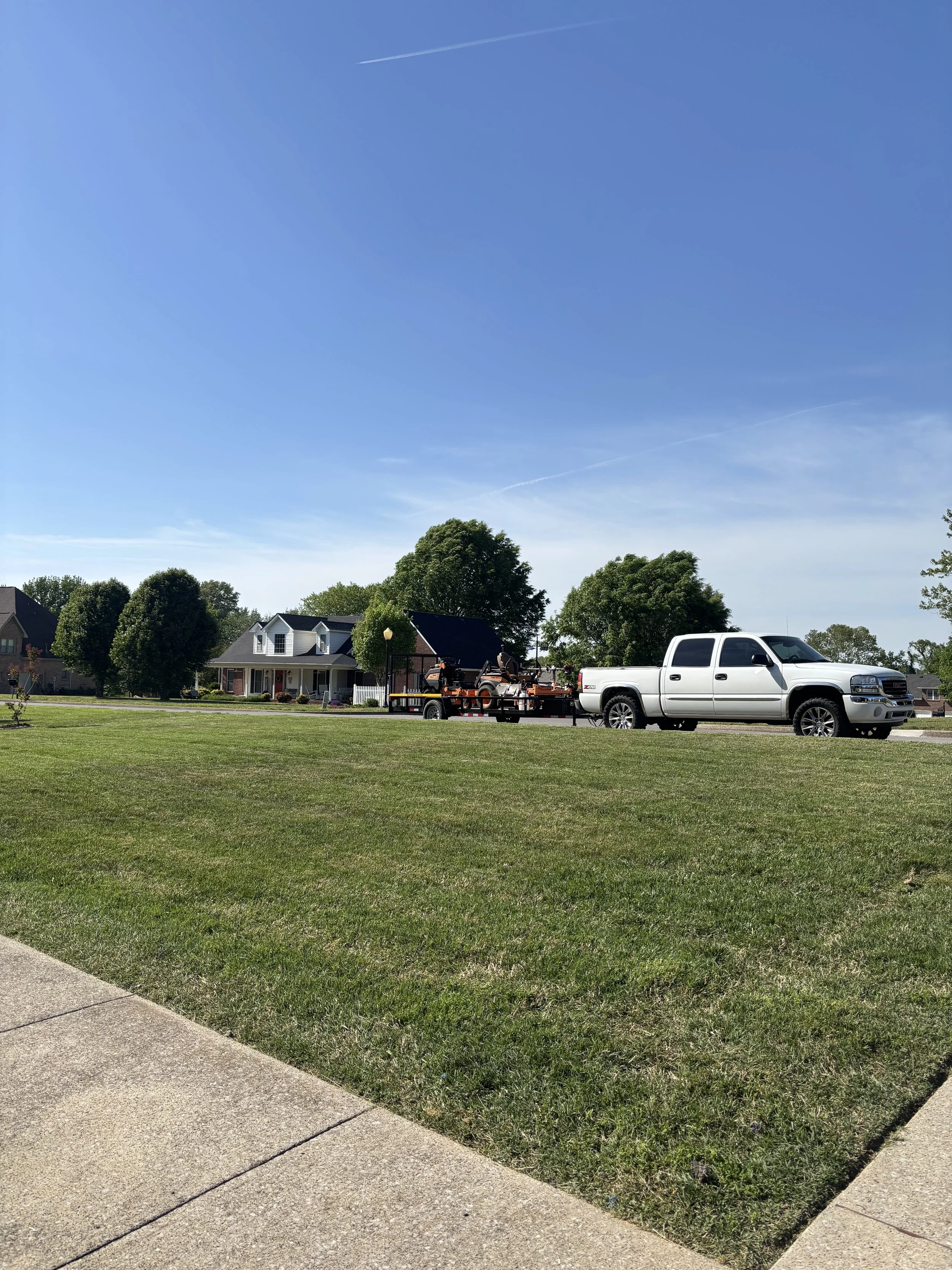 A suburban neighborhood with houses, trees, and a white pickup truck towing lawn equipment on a trailer on a sunny day.