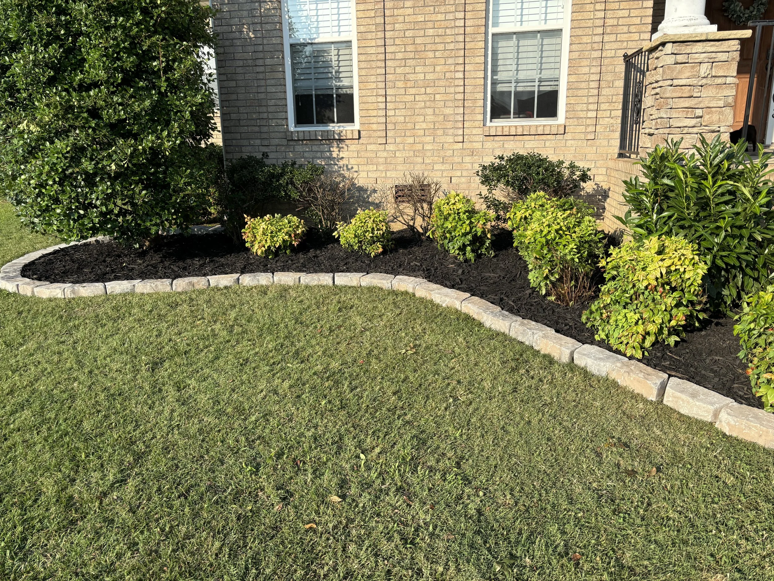 Landscaped garden bed with shrubs and plants, bordered by stone bricks, in front of a brick house with two windows and a porch with railing.
