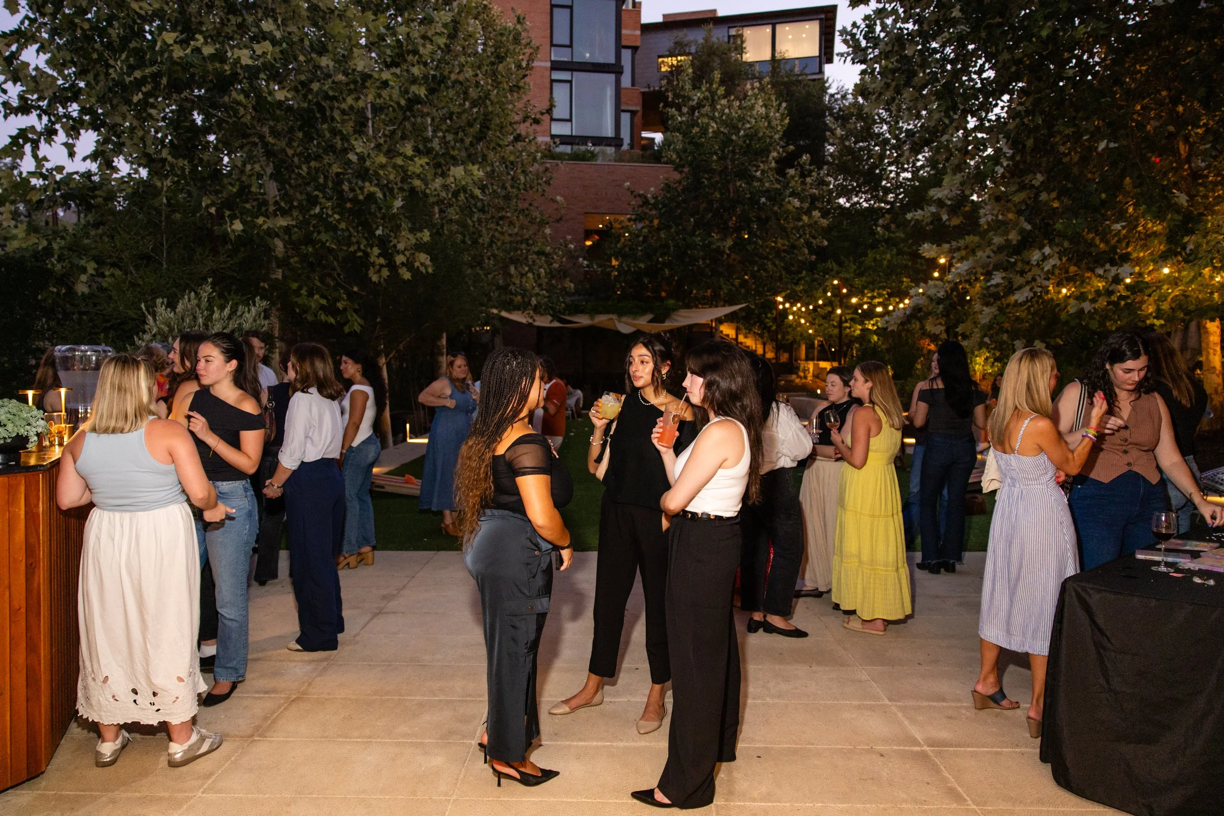 People socializing at an outdoor gathering during the evening, with string lights hung across trees and buildings in the background, and drinks in hand.
