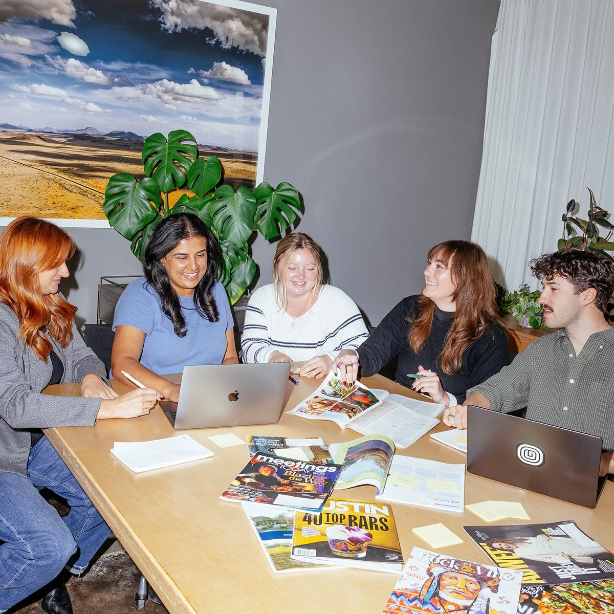 Group of five people sitting around a table, engaged in a discussion, with magazines, notebooks, and laptops in front of them. A large plant and a landscape photograph are visible in the background.
