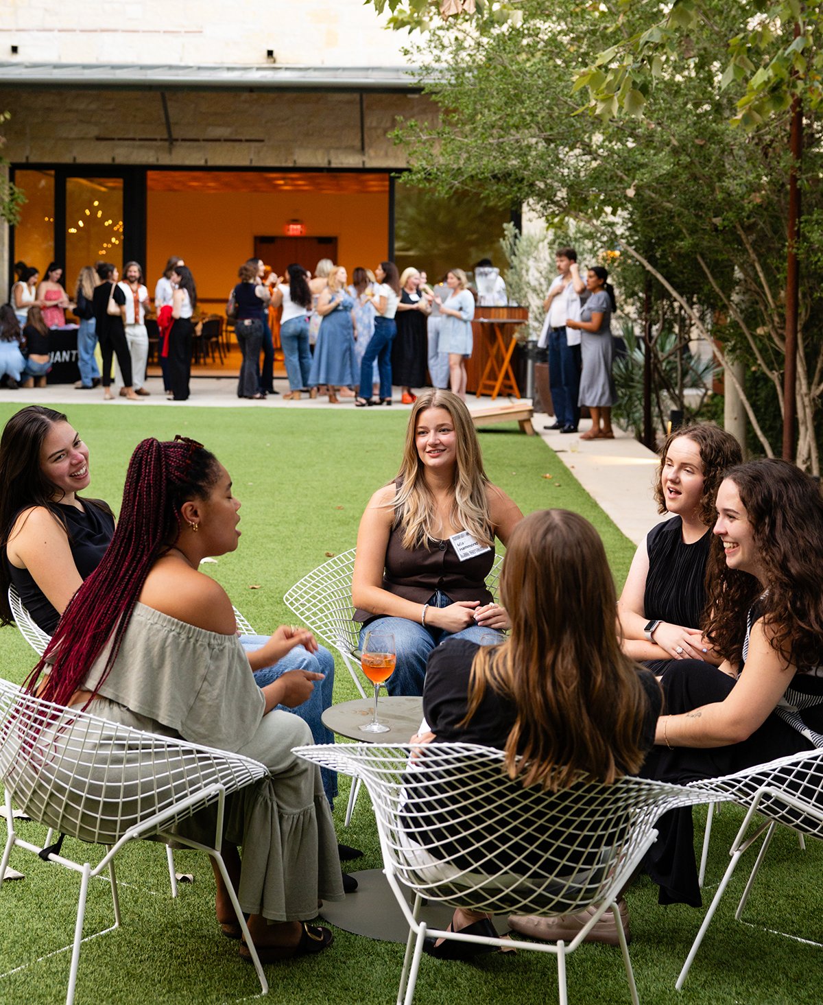 Group of women sitting on white wire chairs, chatting and laughing at an outdoor social event with a green lawn and a building with people in the background.