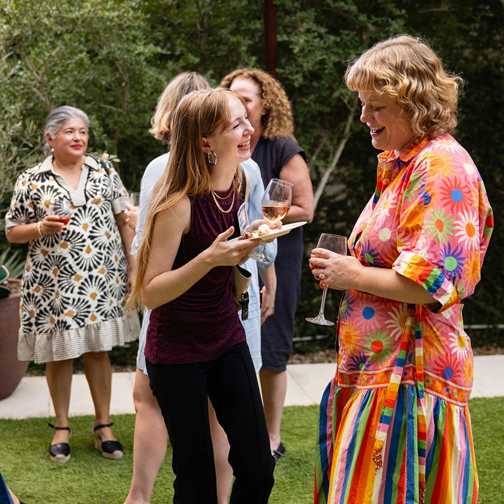 Women laughing and talking at an outdoor gathering, holding wine glasses, with a woman in colorful dress and others in casual attire, on a green lawn with trees in the background.